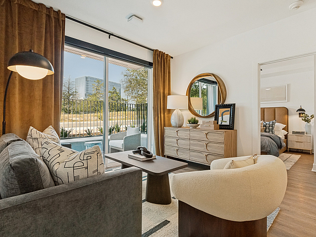 Bright and welcoming living room with stylish decor, plush seating, and a view of the outdoor greenery through large glass doors, complemented by an elegant bedroom entrance.