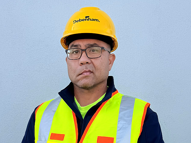 Confident construction worker in a high-visibility vest and safety helmet standing against a smooth gray wall.
