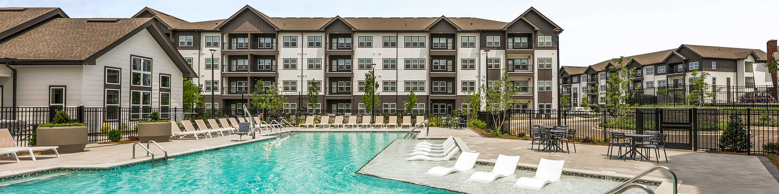 Luxury apartment complex featuring a sparkling swimming pool with elegant lounge chairs, surrounded by modern multi-story buildings and lush landscaping.