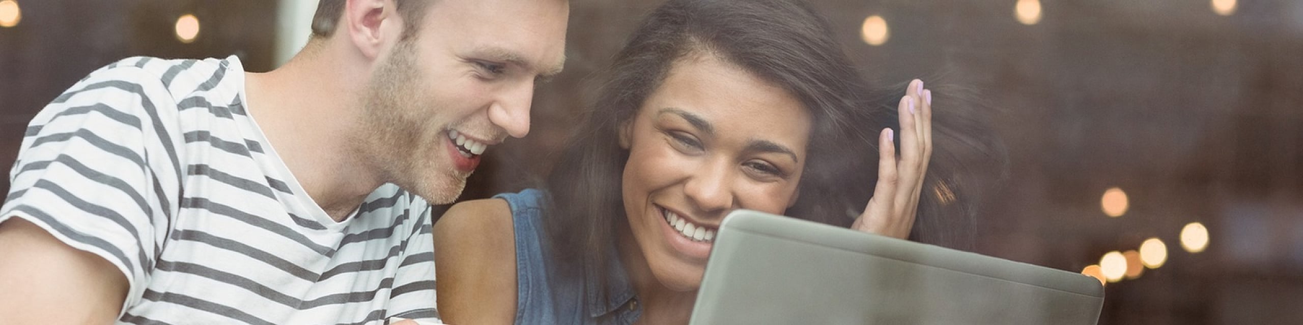 Joyful couple sitting at a caf&eacute; window, sharing a laptop and coffee, creating a warm and inviting atmosphere.