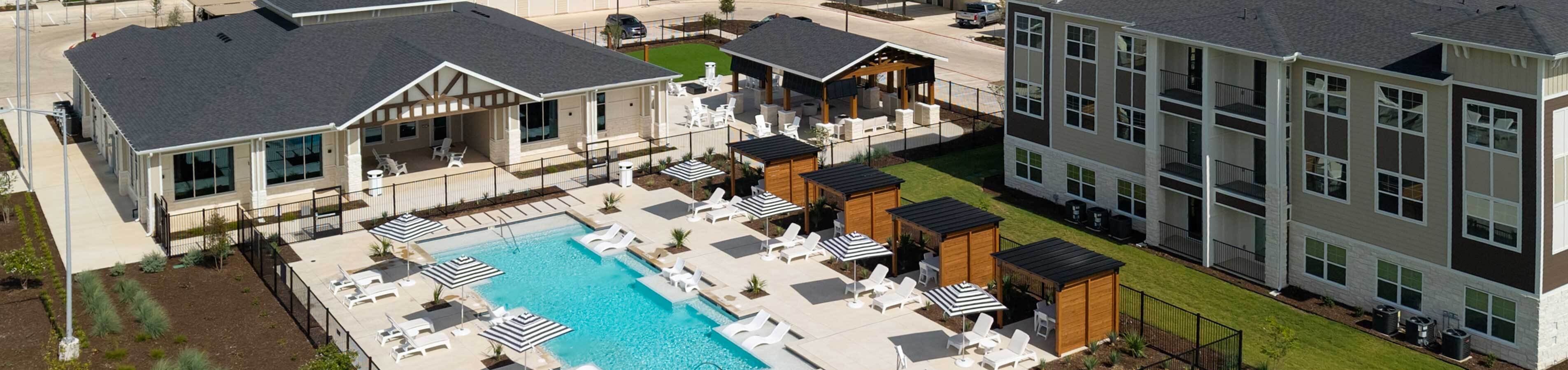 Aerial view of a modern apartment complex featuring a large resort-style swimming pool, striped umbrellas, lounge seating, wooden cabanas, and a community clubhouse alongside multi-story residential buildings.