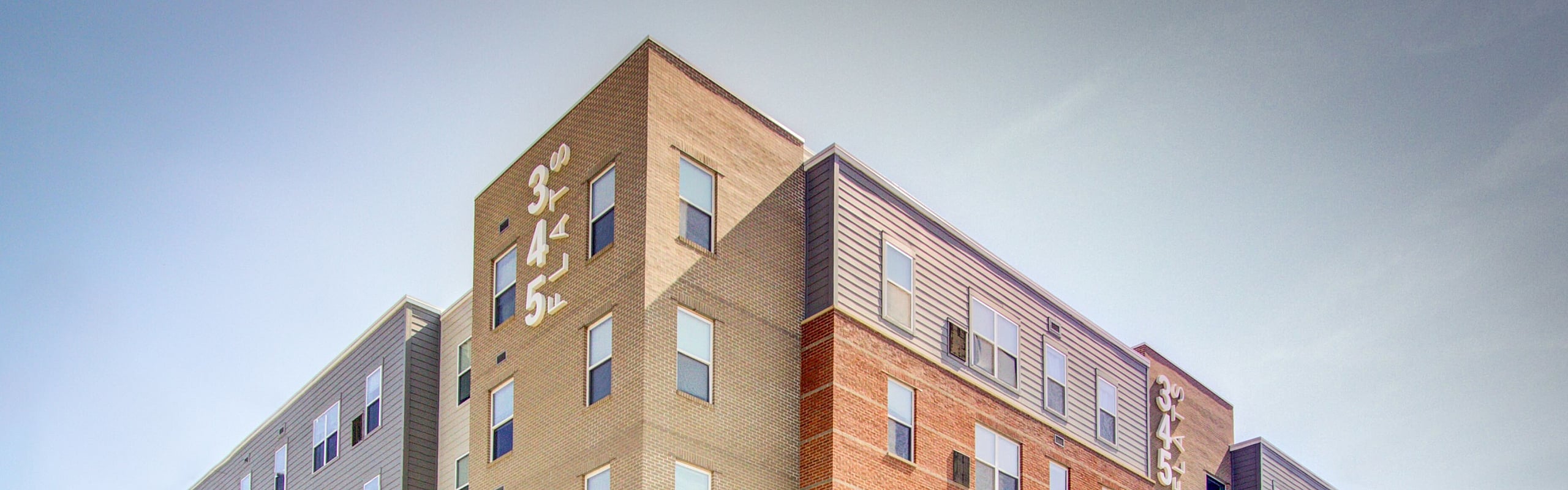 Modern multi-story apartment building with red-brick and neutral-toned facade, featuring large windows and a lush green entrance area.