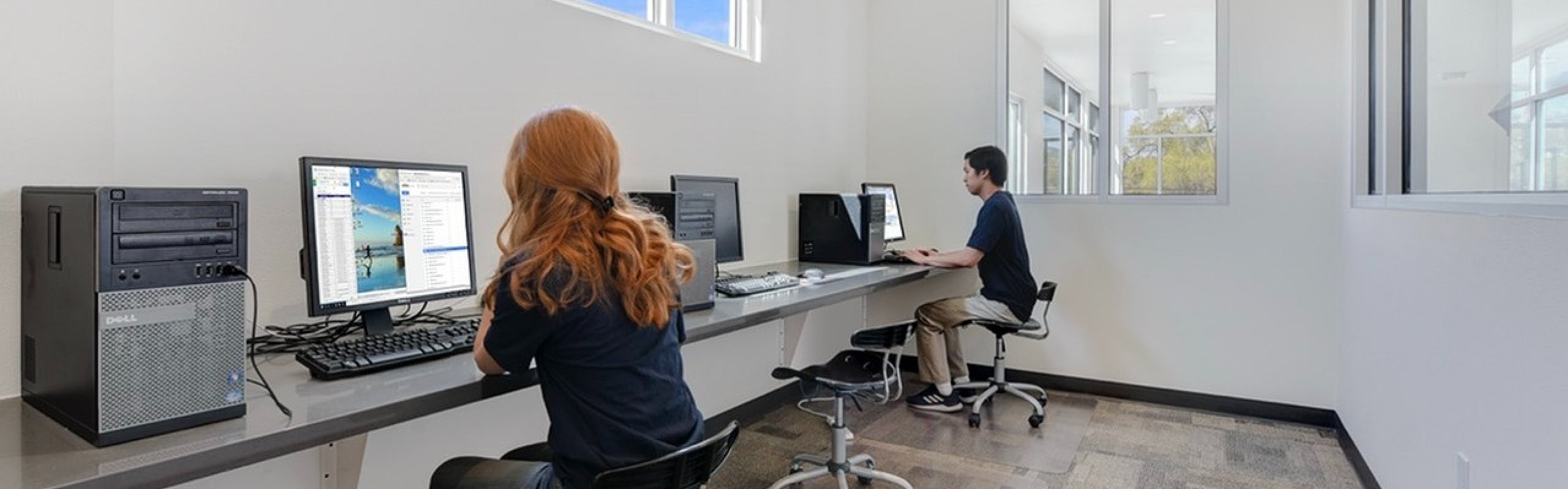 Bright office space with modern workstations and natural light, featuring two people focused on their work at sleek computer desks.