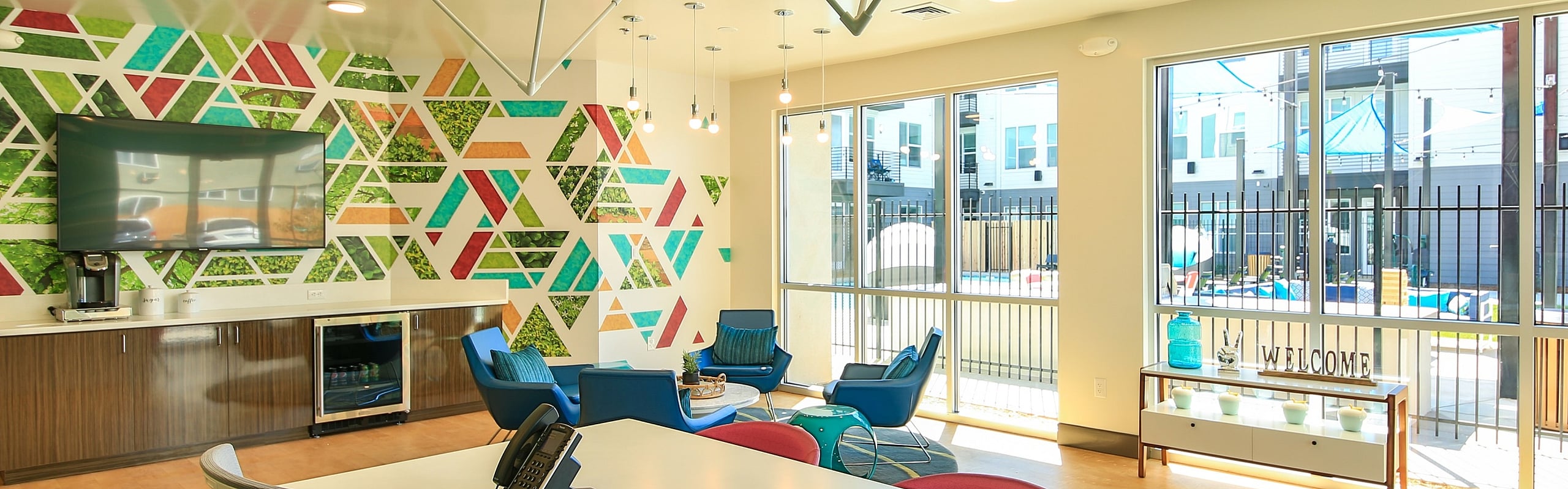 Bright and modern apartment lounge with geometric wall art, contemporary lighting, and cozy seating by large sunlit windows.