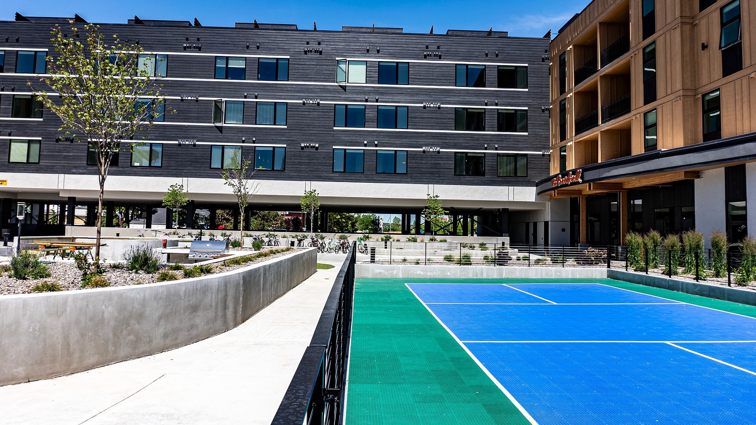 Modern apartment complex with sleek architecture and vibrant outdoor tennis court, surrounded by landscaped pathways.