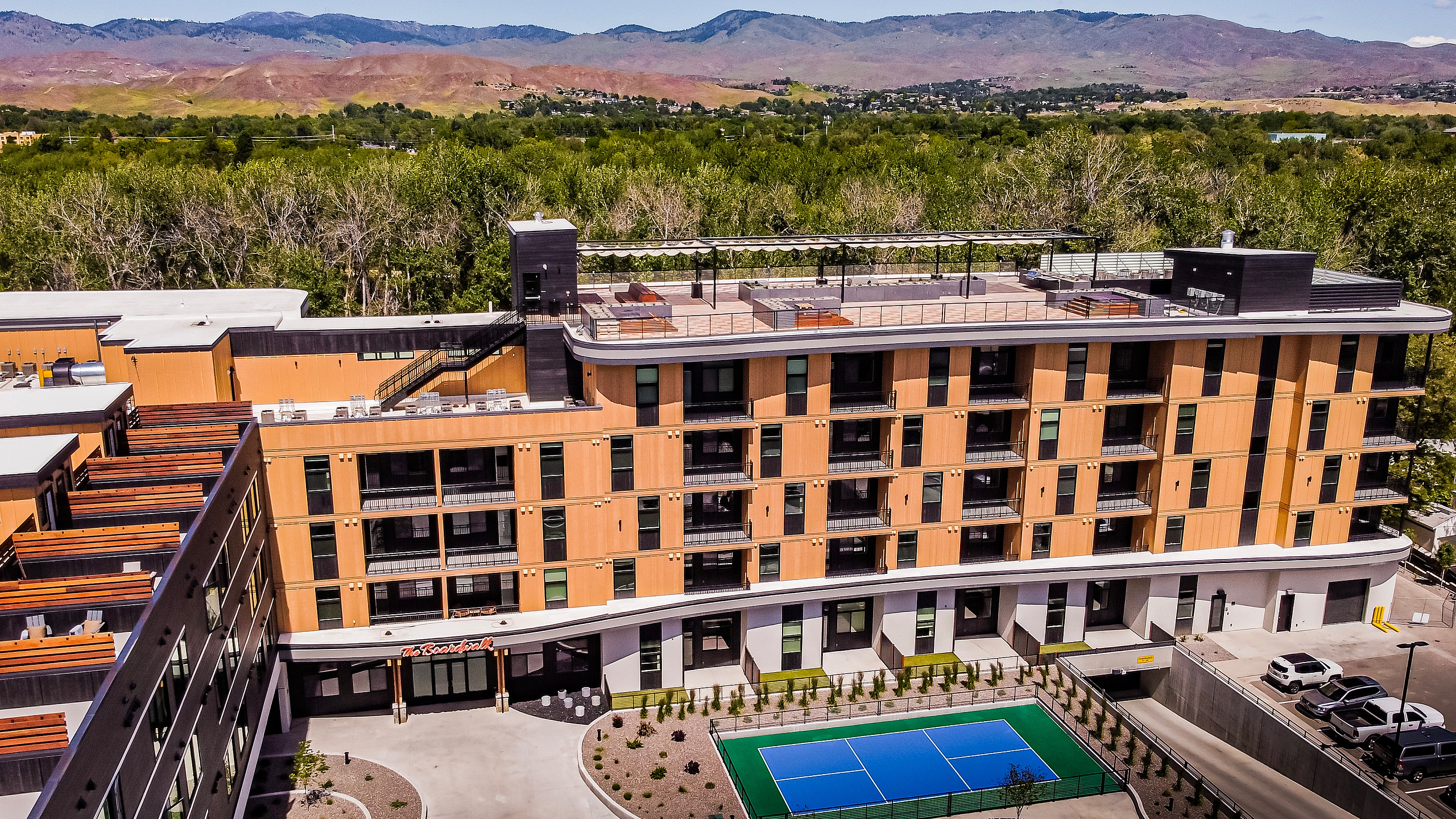 Modern apartment building with striking wood accents and ample balconies, featuring a rooftop terrace and a vibrant blue sports court surrounded by lush greenery and scenic mountain views.