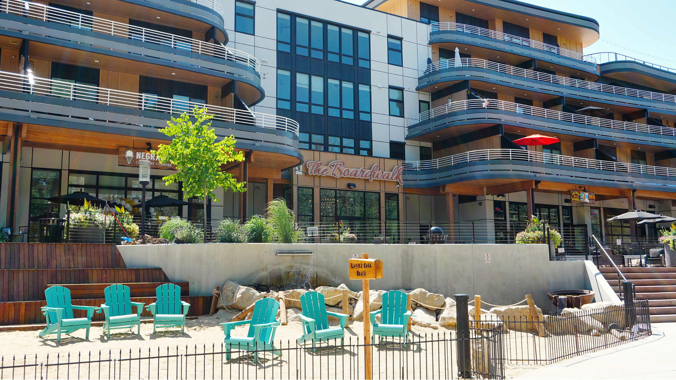 Modern apartment complex with distinctive balconies, ground-level cafe, and vibrant teal outdoor chairs, surrounded by lush greenery and a relaxing ambiance.