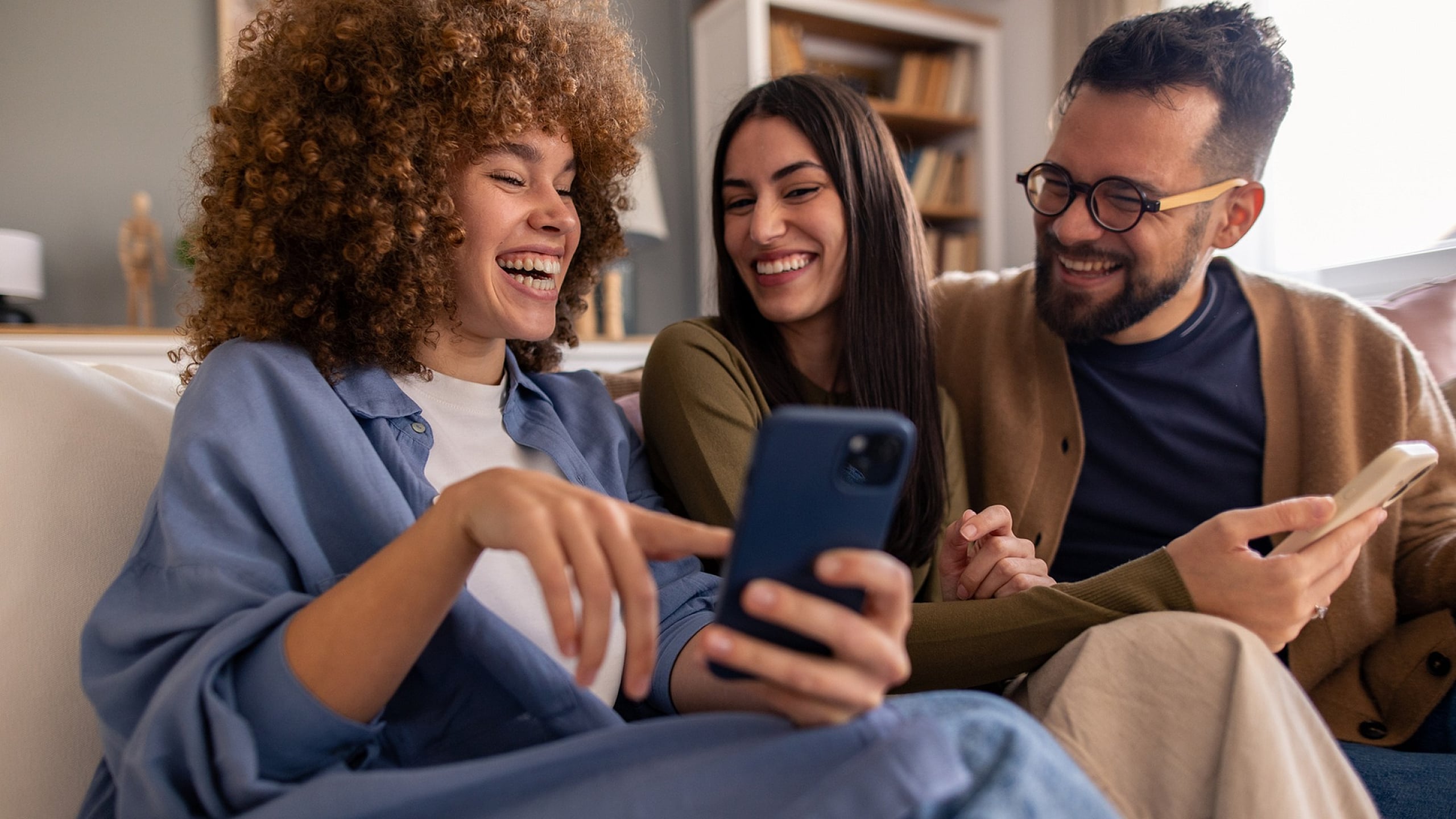 Joyful friends enjoying a cozy living space, sharing laughs and moments on their smartphones.