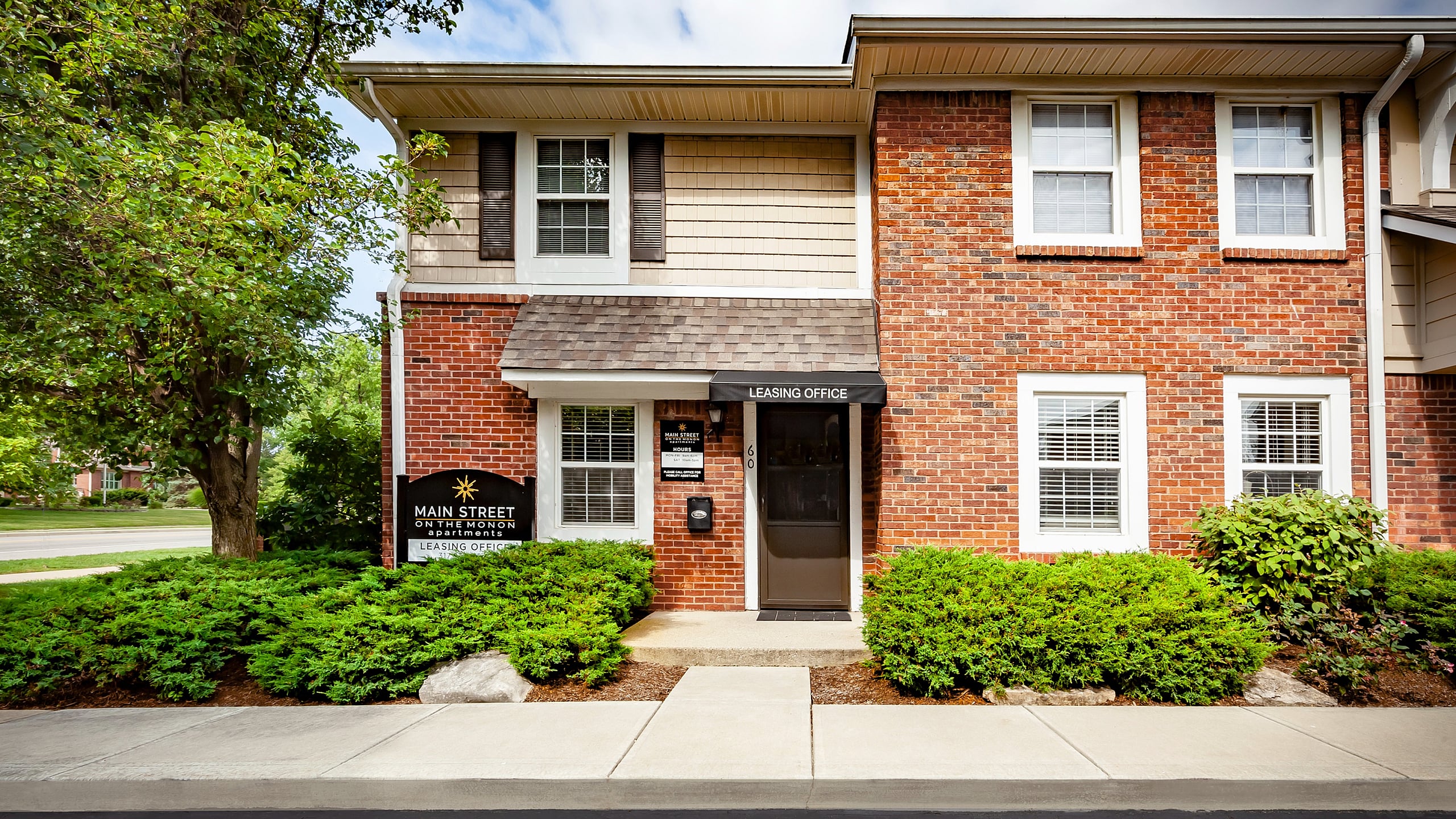 Charming brick leasing office at Main Street on the Monon Apartments, surrounded by lush greenery under a bright blue sky.