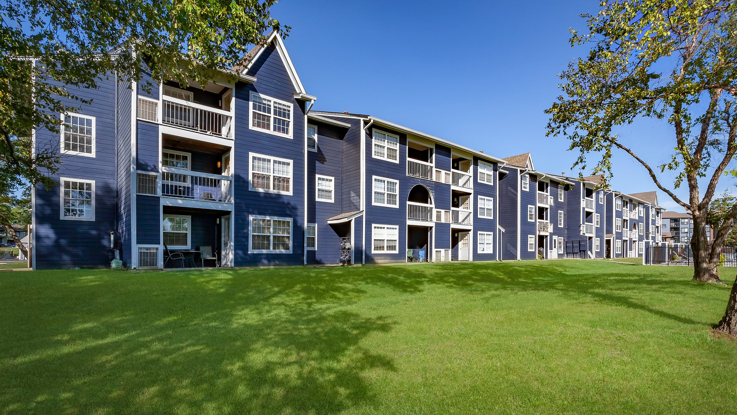 Charming blue apartment building with bright white accents, surrounded by lush green lawns and tall trees, under a clear blue sky.