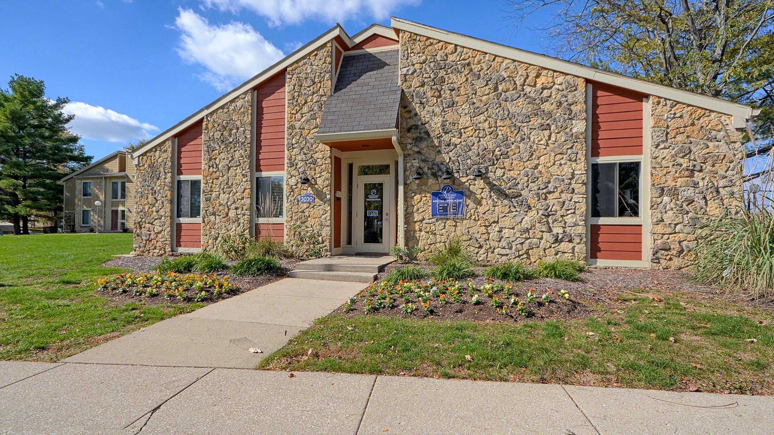 Charming stone facade building with an inviting entrance, surrounded by lush greenery and vibrant flowerbeds under a clear blue sky.