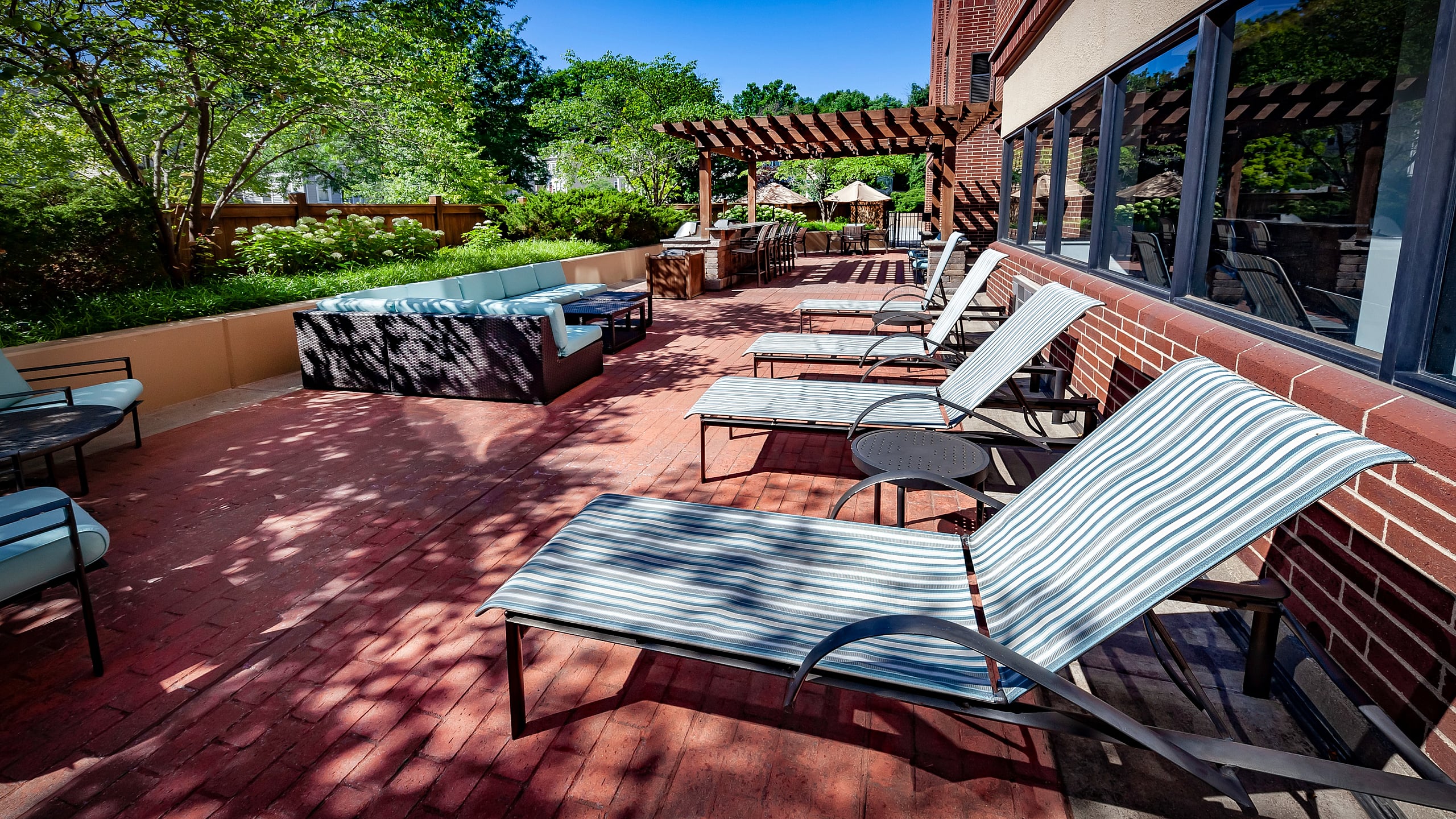 Sunny outdoor patio with stylish lounge chairs and cozy seating area under a wooden pergola, surrounded by lush greenery.