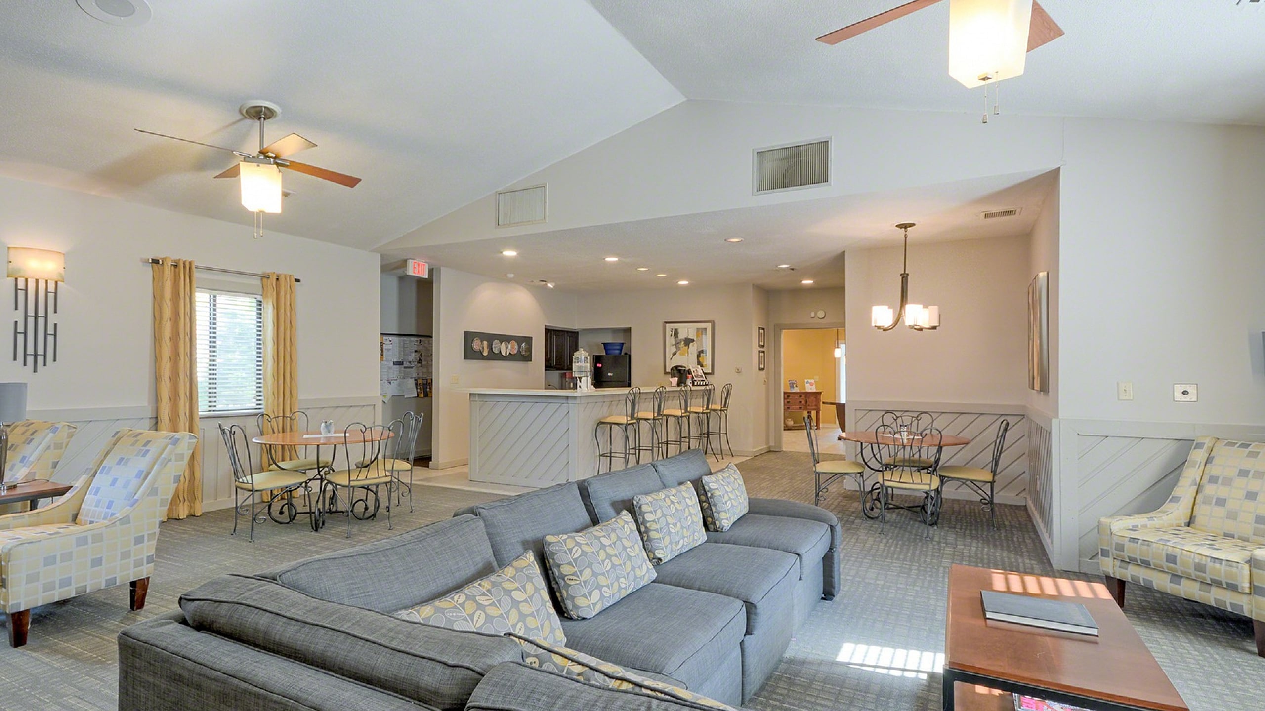 Bright and welcoming apartment lounge featuring stylish gray sectional seating, modern ceiling fans, and a cozy dining area with elegant light fixtures.