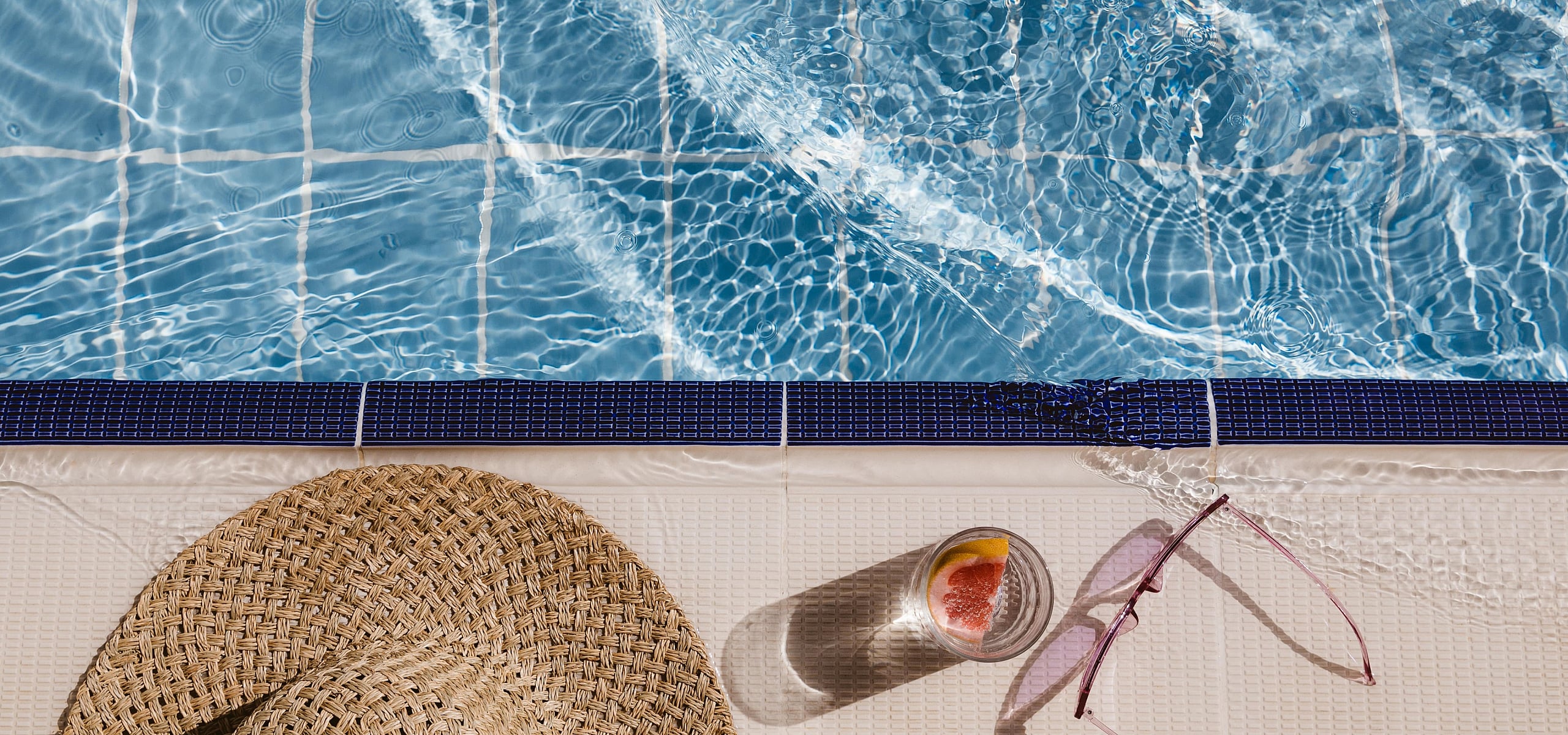 Inviting poolside escape featuring sparkling blue water, a woven sunhat, chic sunglasses, and a refreshing fruit beverage.