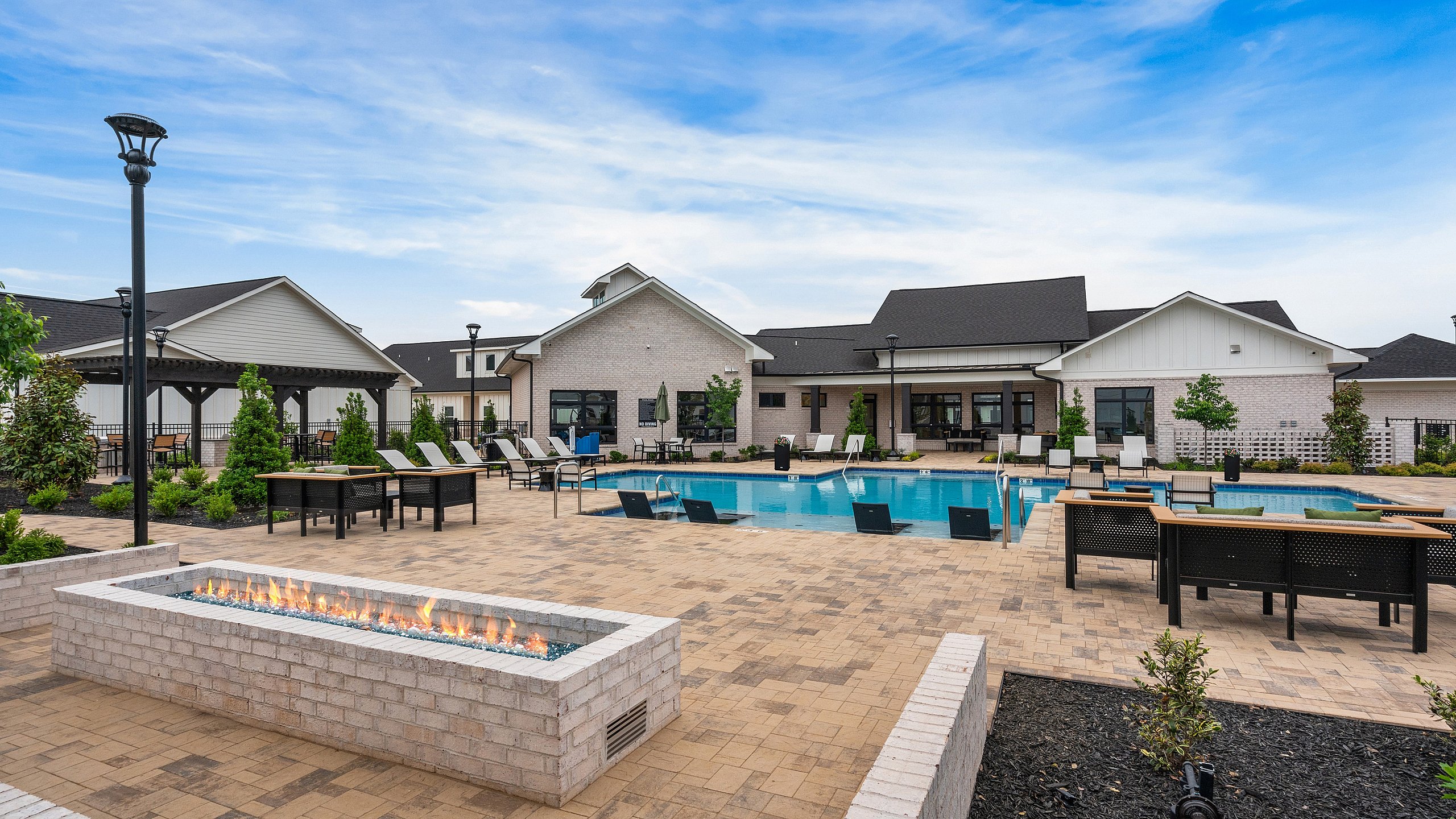 Luxurious apartment complex featuring a sparkling pool, modern lounge chairs, and an elegant outdoor fire pit area against a serene blue sky backdrop.