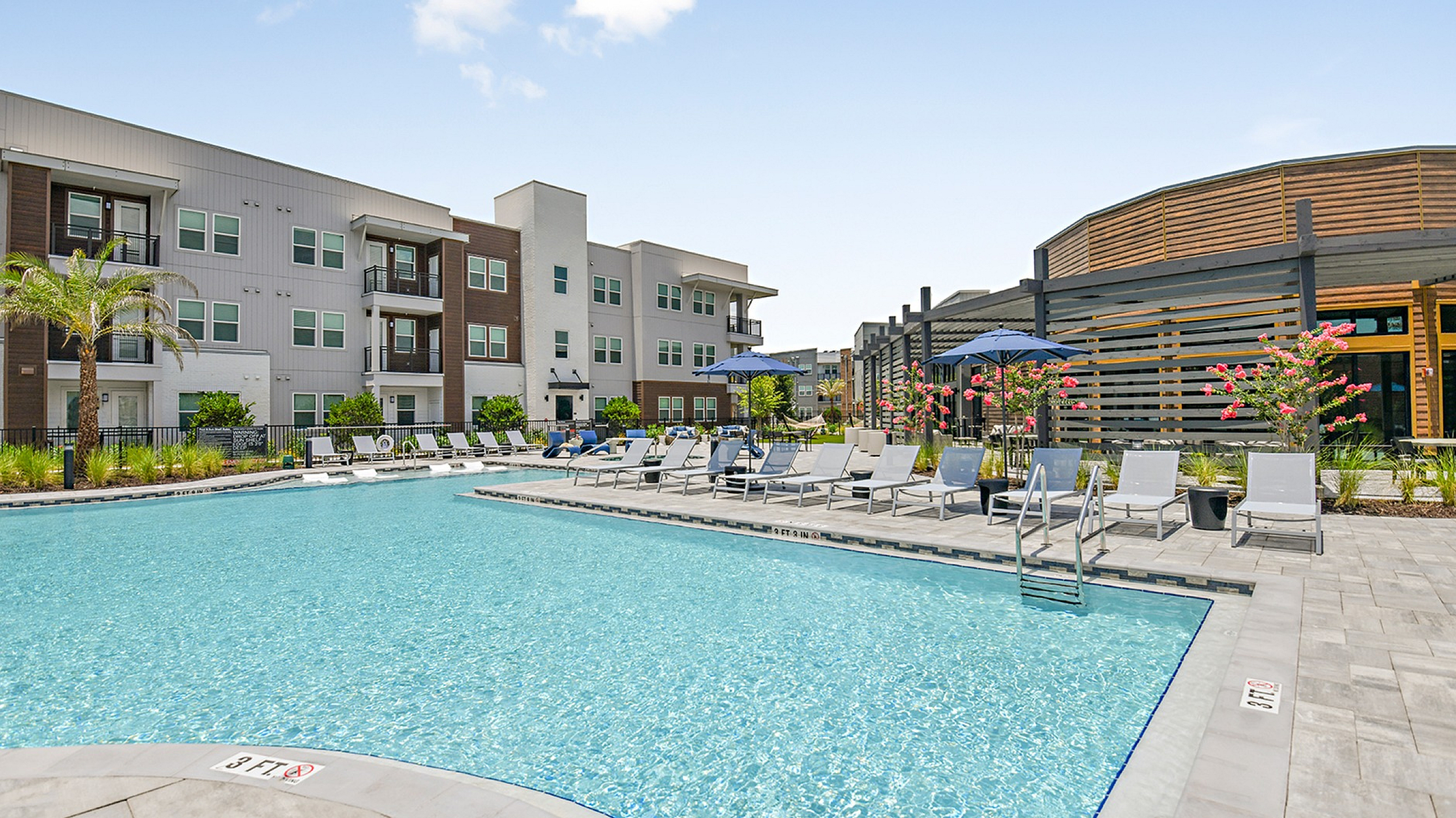 Luxurious apartment complex with a sparkling pool, stylish loungers, and vibrant landscaping under a bright blue sky.