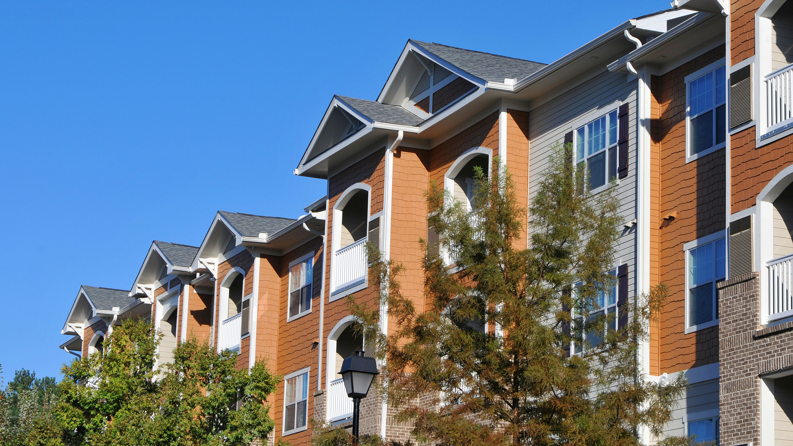 Charming, modern apartment complex with elegant architecture and sunlit balconies, surrounded by mature trees and blue skies.