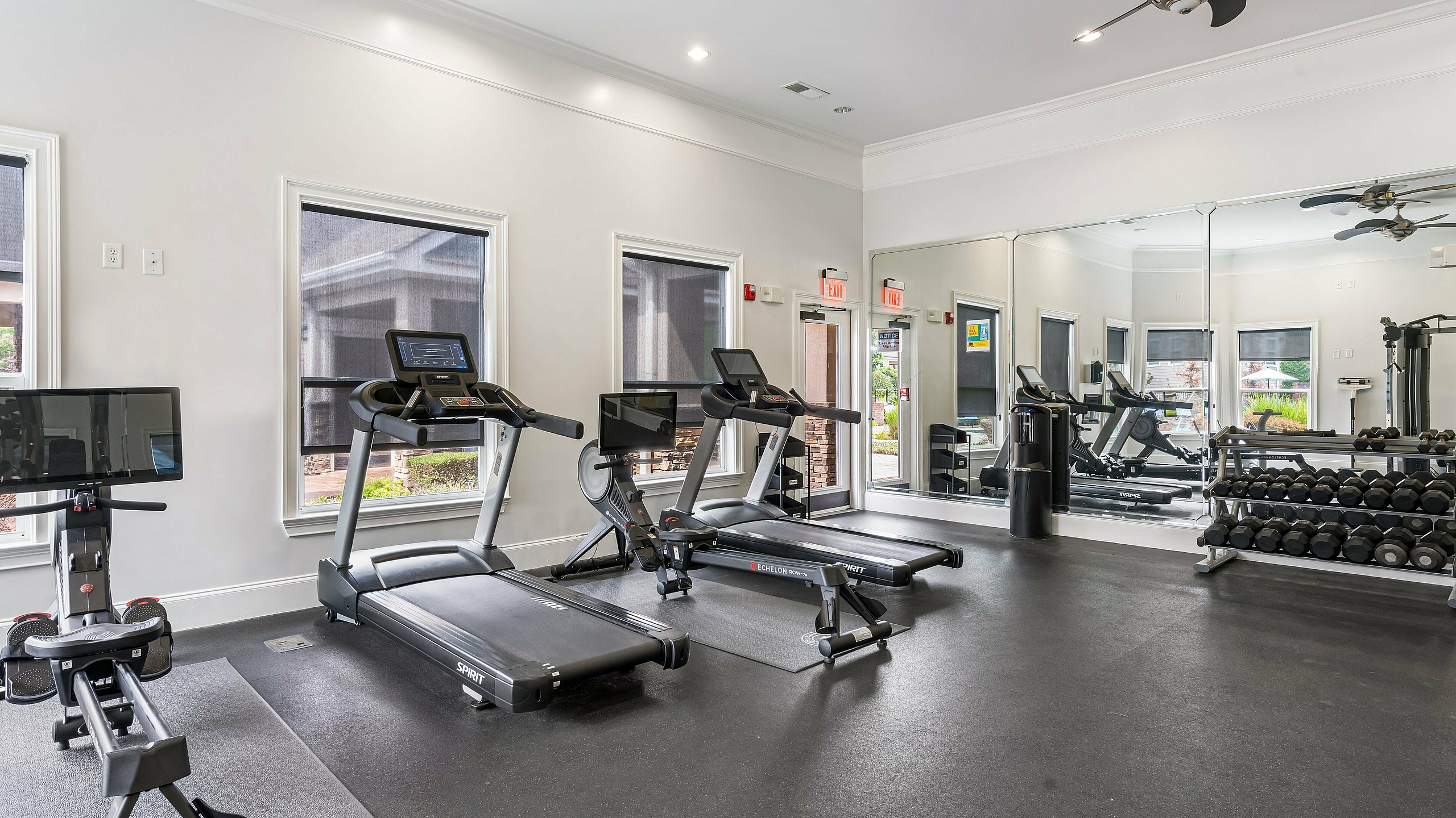 Modern gym area with state-of-the-art equipment, including treadmills, a rowing machine, and a full set of free weights, enhanced by natural light and large mirrors.