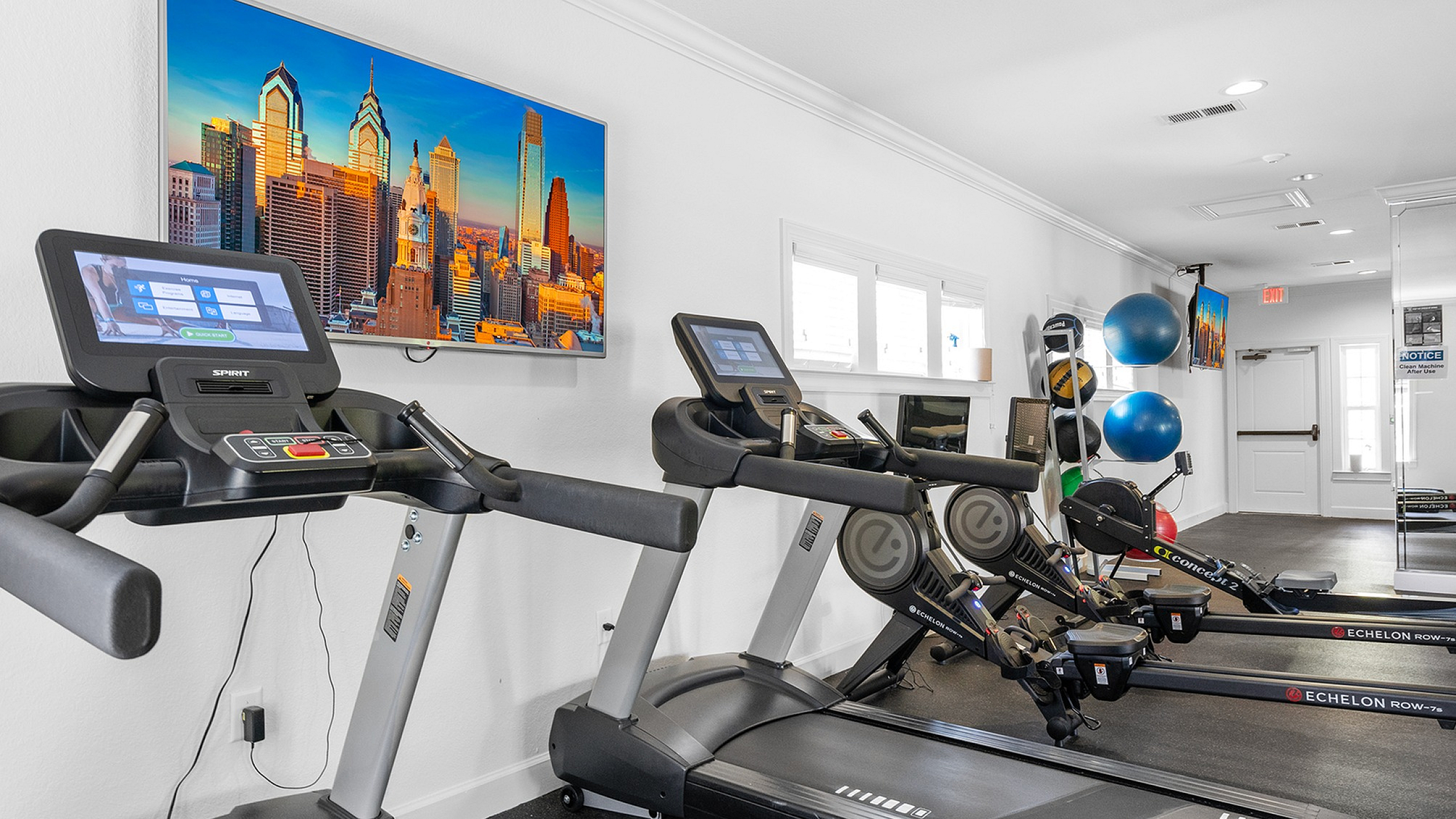Modern apartment gym featuring state-of-the-art treadmills, rowing machines, and a vibrant city skyline photograph.