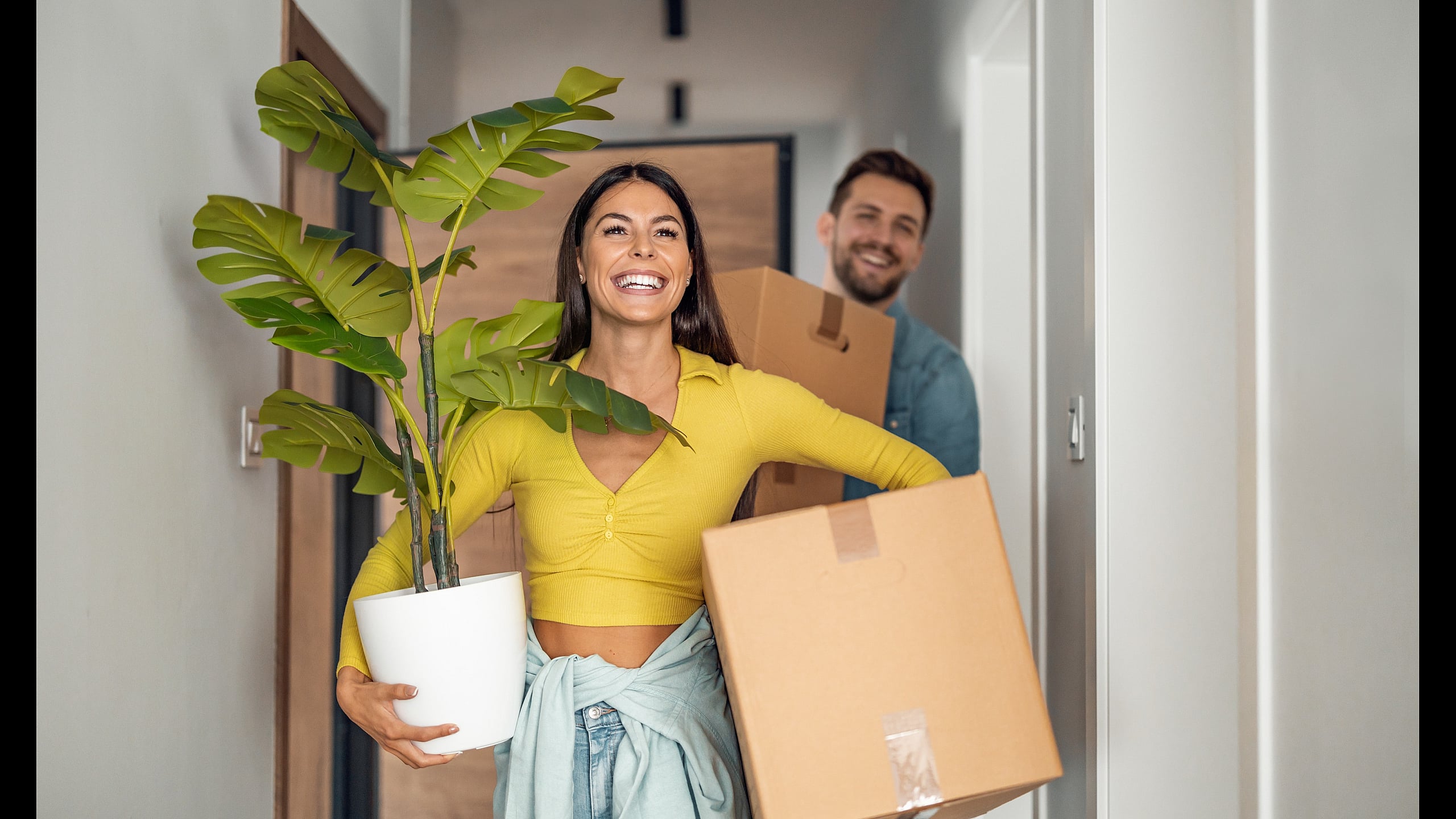 Joyful couple moving into a bright new apartment, carrying a potted plant and boxes, symbolizing a fresh start.