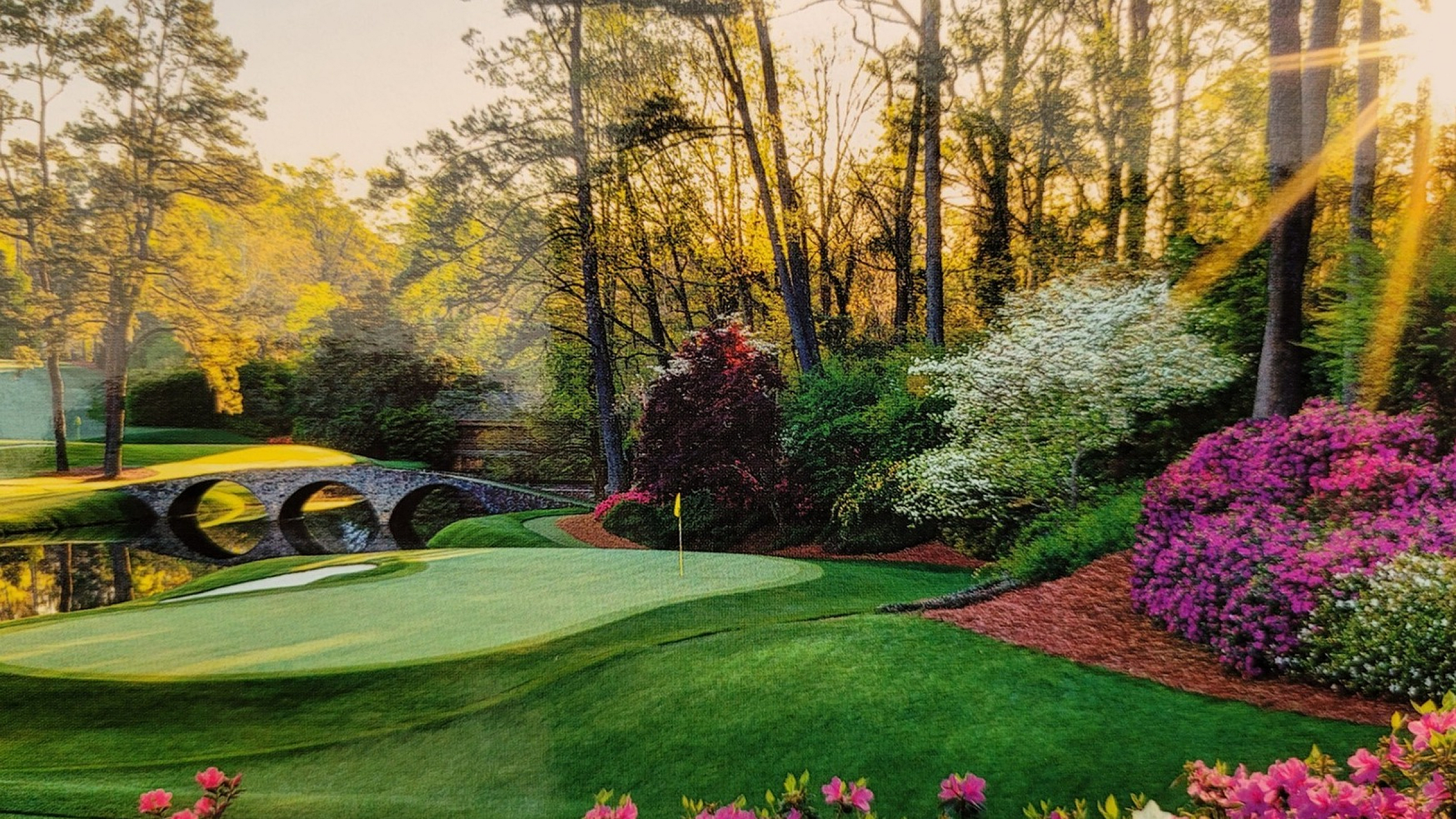 Picturesque landscape with vibrant flowers, a serene pond, and a stone bridge bathed in golden sunlight.