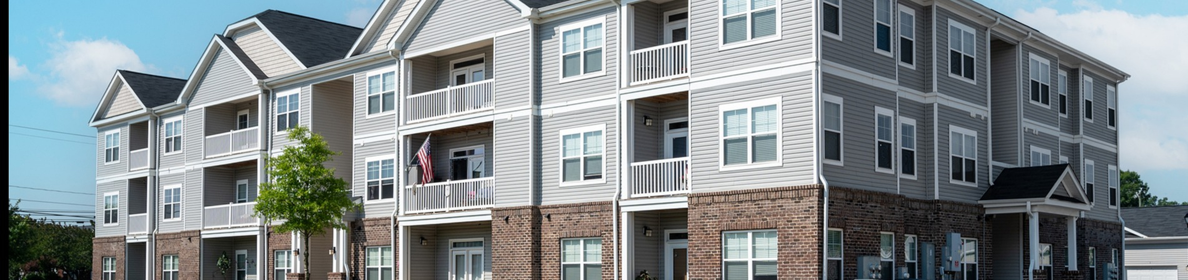 Modern three-story apartment building with elegant balconies and attractive landscaping under a bright blue sky.