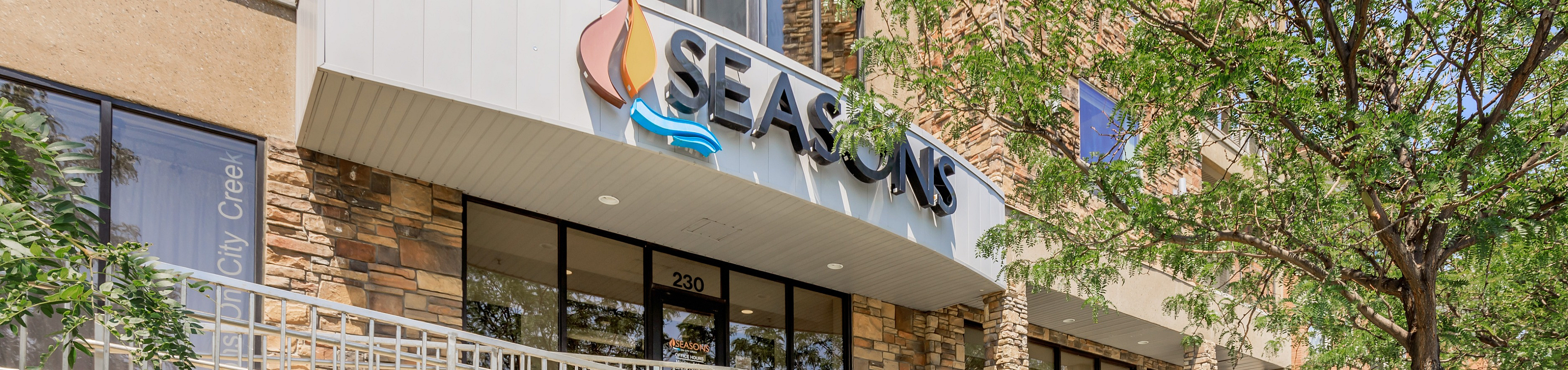 Inviting entrance of a modern apartment building named 'Seasons,' featuring sleek architecture and lush green surroundings.