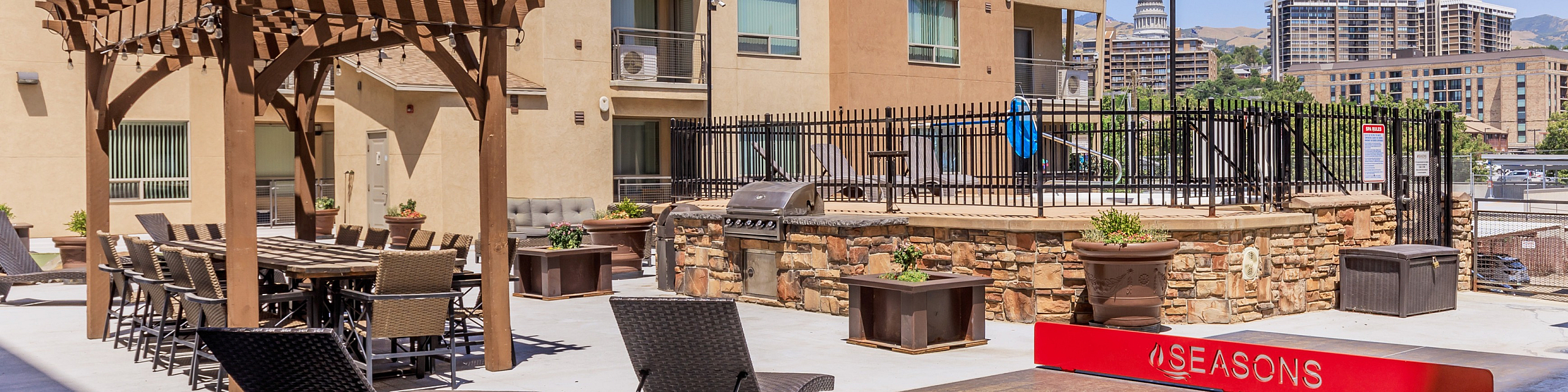 Inviting outdoor courtyard featuring modern lounge chairs, a pergola-shaded dining area, grill station, and a ping pong table, set against a backdrop of contemporary apartment buildings.