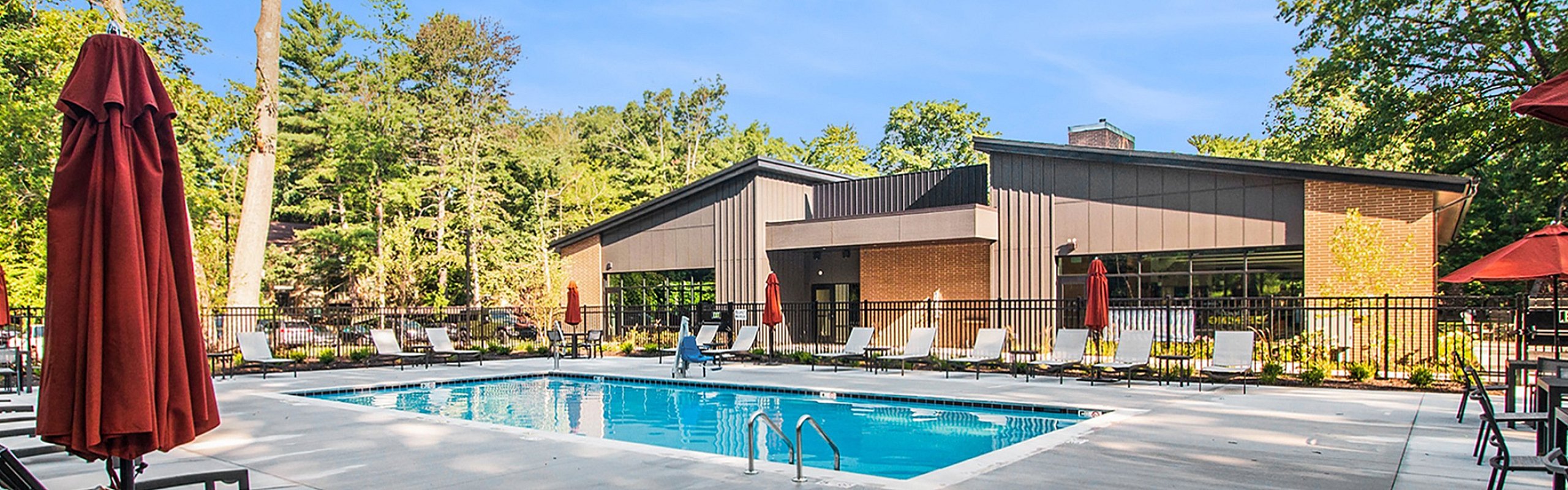 Luxurious apartment complex pool area featuring a pristine swimming pool, modern lounge chairs, and vibrant red umbrellas, surrounded by lush greenery and contemporary architecture.