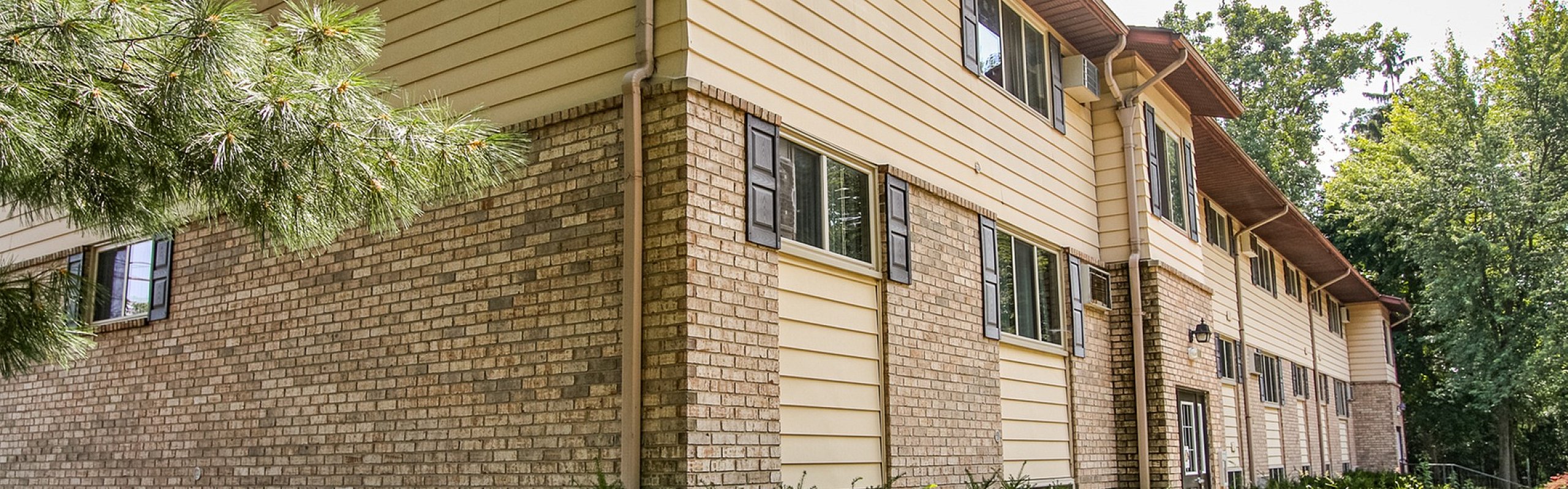 Charming apartment building showcasing elegant brick and siding exterior with lush green surroundings, perfect for serene living.