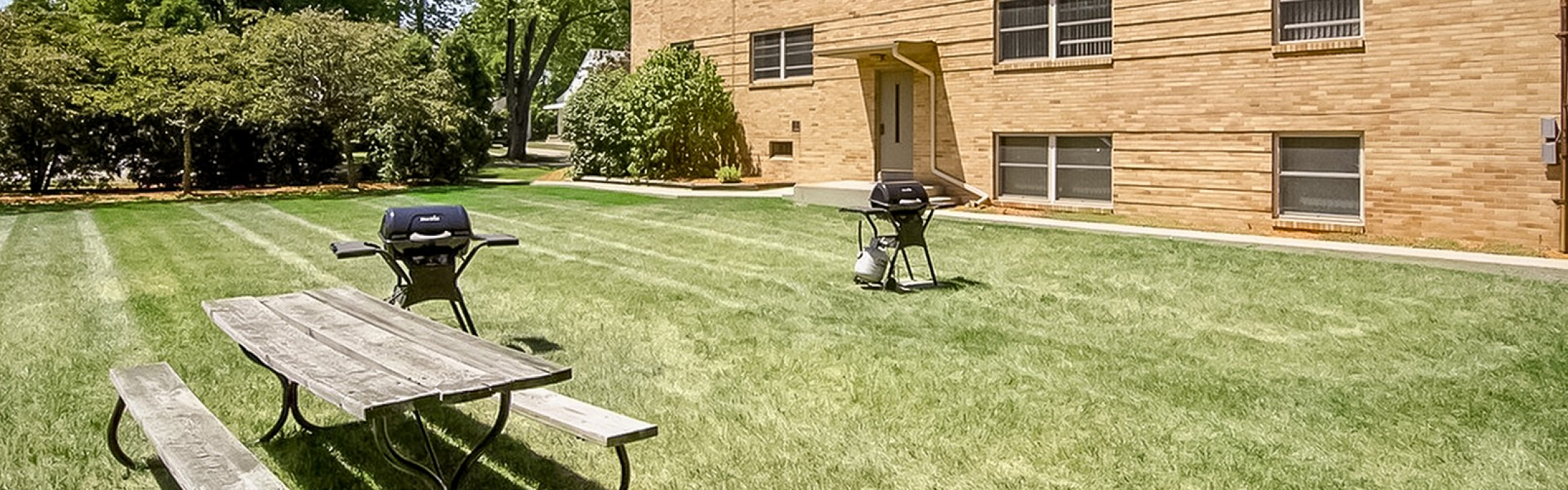 Charming apartment exterior with lush green lawn, outdoor seating, and grills, perfect for sunny gatherings.