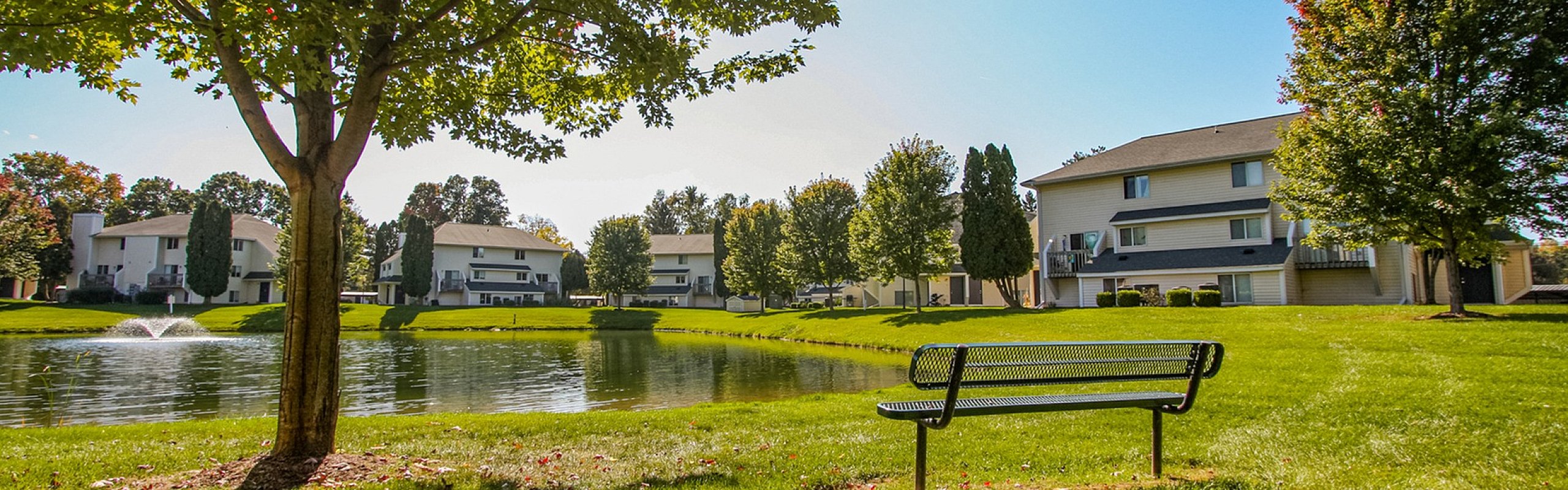Serene apartment complex surrounded by lush greenery, featuring a tranquil pond and charming outdoor seating under a sunlit sky.