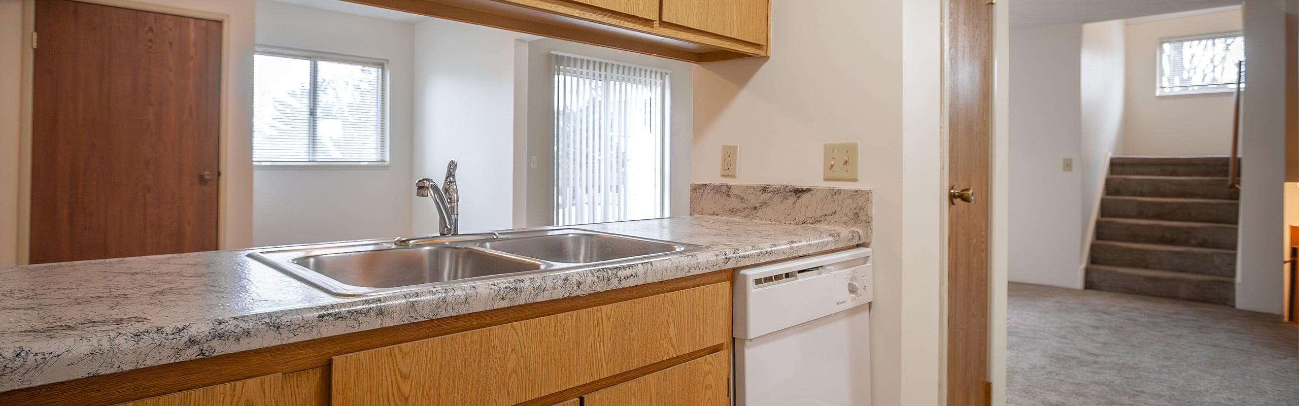 Sleek kitchen with marble countertop and modern appliances, adjacent to a carpeted living area with natural light and staircase.
