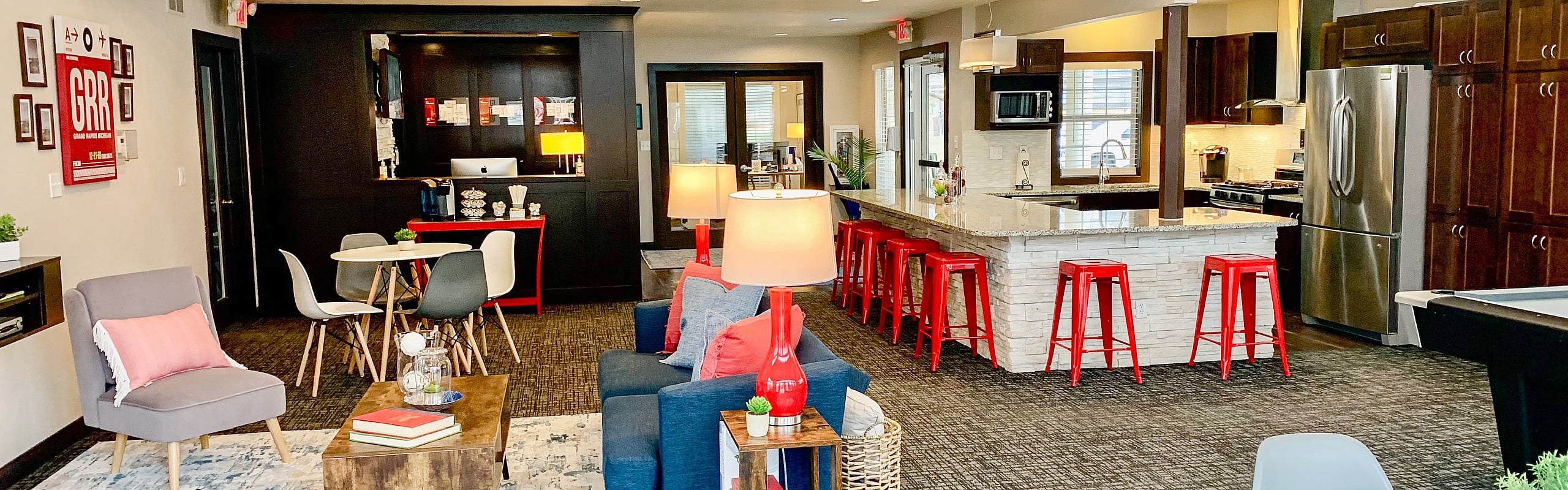 Inviting apartment community lounge with modern decor, featuring a sleek kitchen with stainless steel appliances and vibrant red bar stools, complemented by cozy seating and warm lighting.