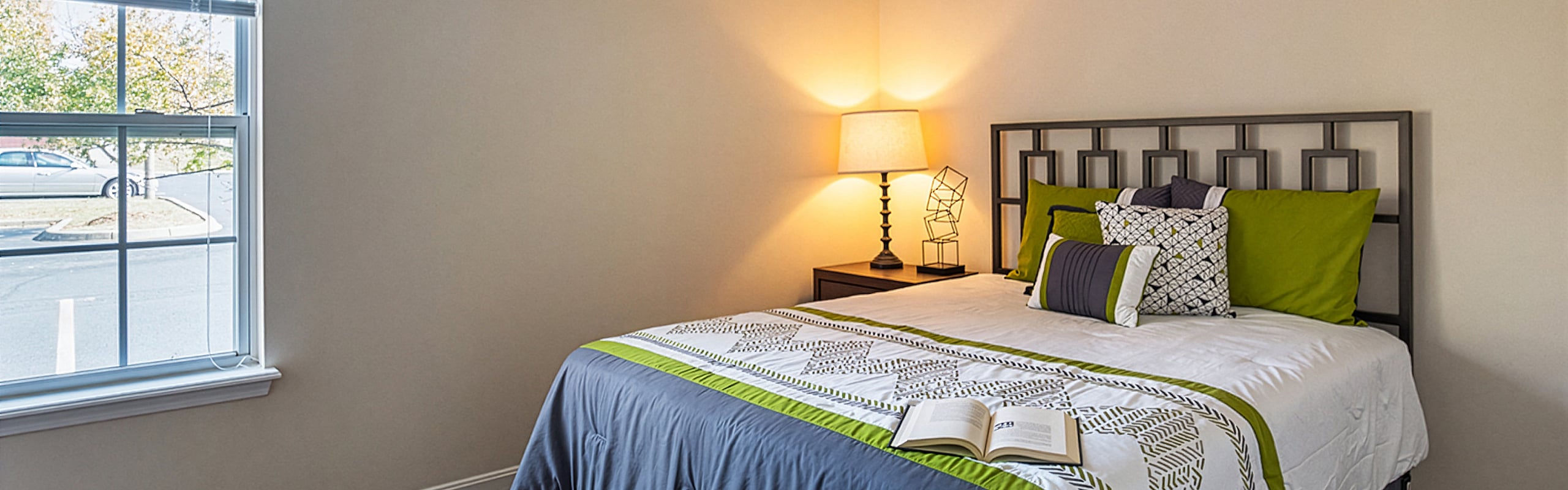 Bright bedroom with elegant decor, featuring a cozy bed with green accents, a stylish lamp, and a large sunlit window.