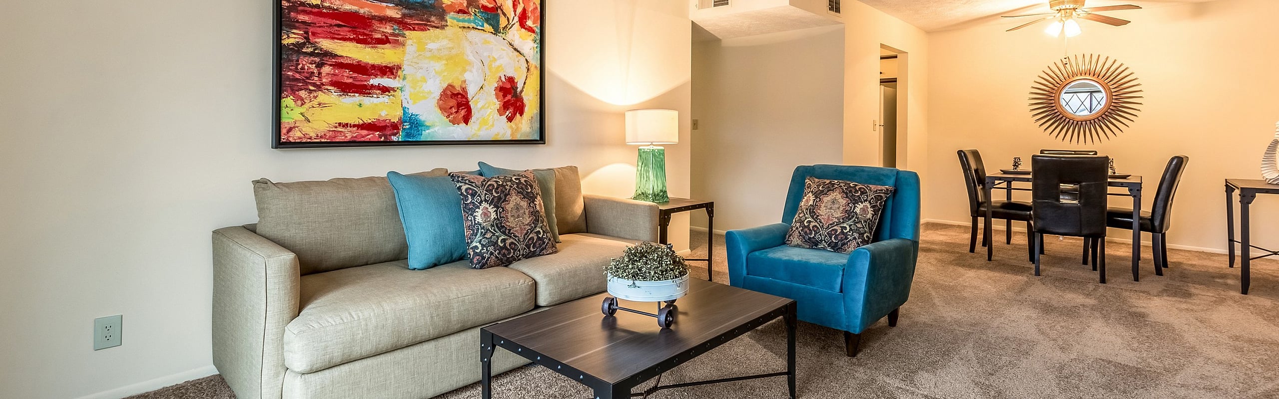 Charming living room with vibrant artwork, cozy beige sofa, and a striking blue armchair, leading to a stylish dining area with a sunburst mirror.