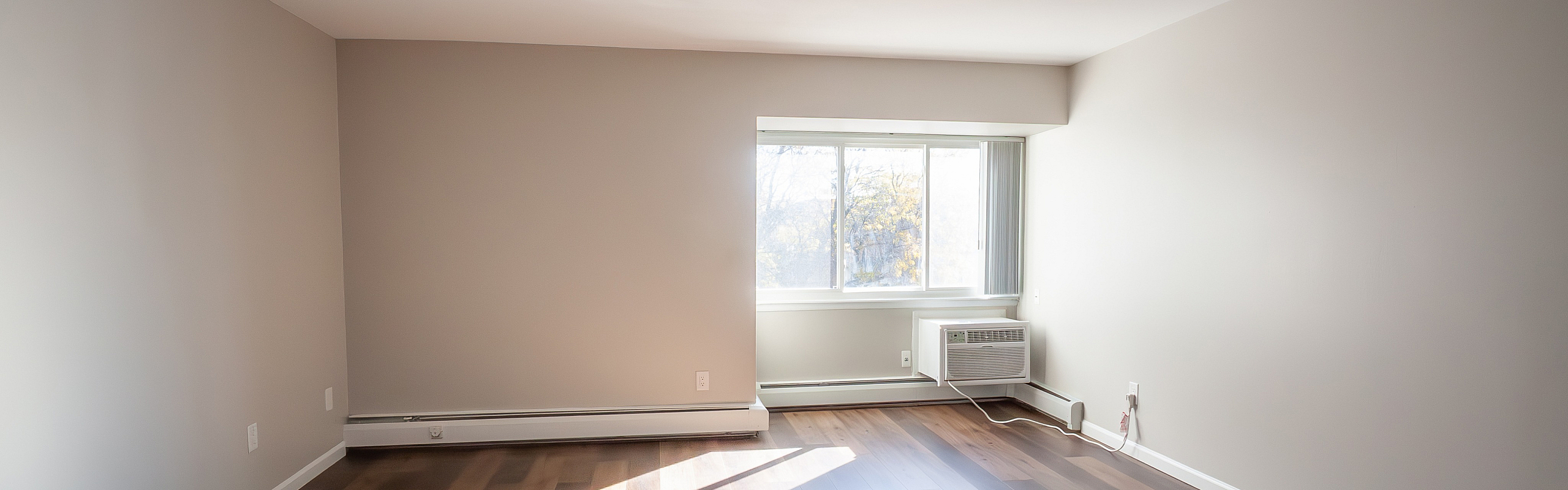 Bright and inviting room with natural light streaming through a large window, highlighting elegant wood flooring and a built-in air conditioner.