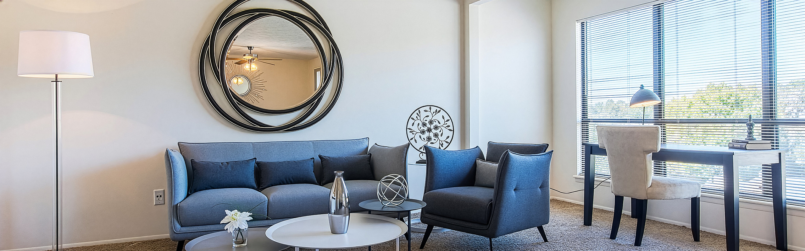 Elegant living room with modern blue furniture, a stylish circular mirror, and a cozy workspace by large windows.