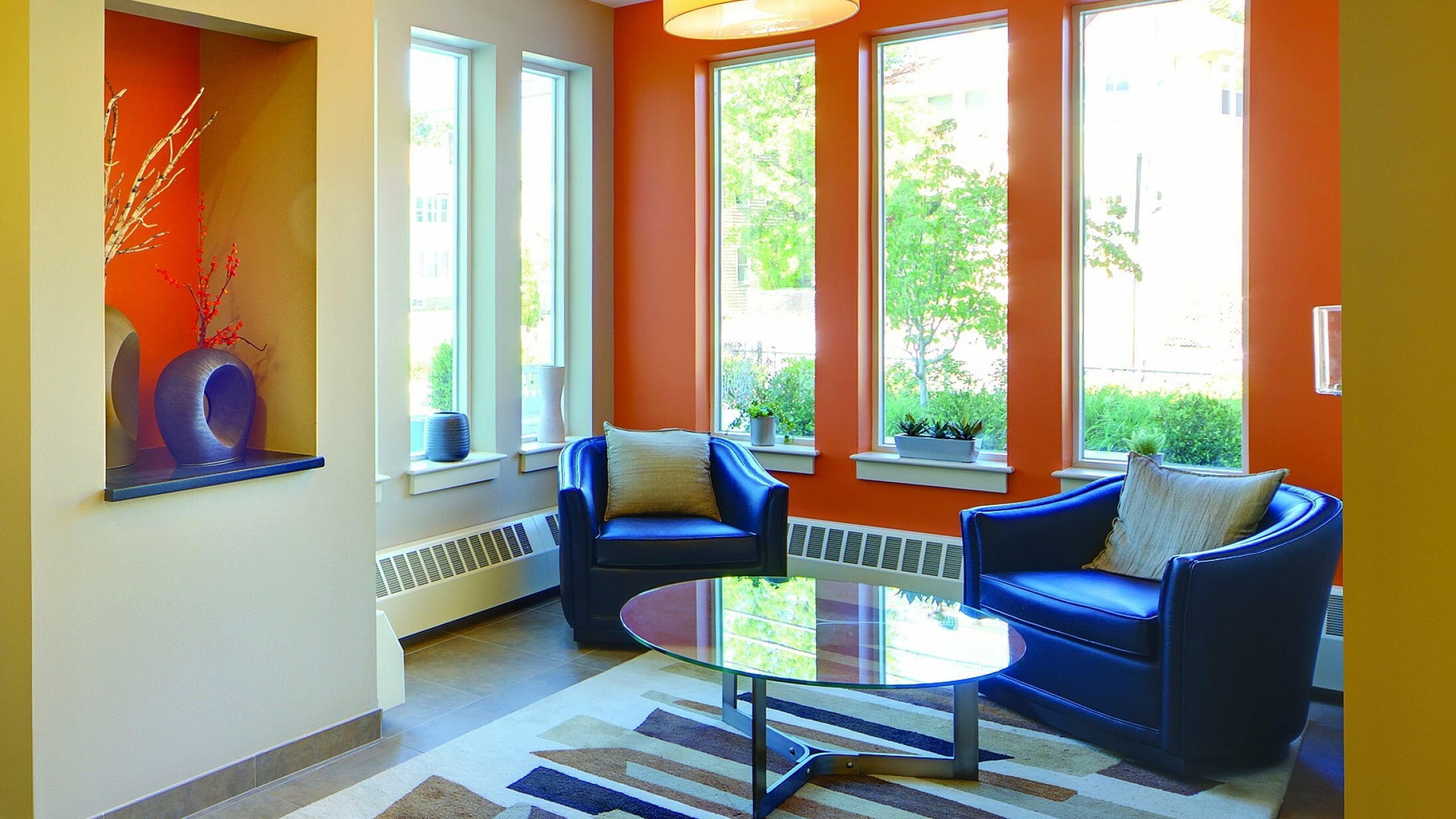 Inviting apartment lobby with modern decor, vibrant orange accent wall, and comfortable seating area featuring blue armchairs and a stylish glass coffee table.