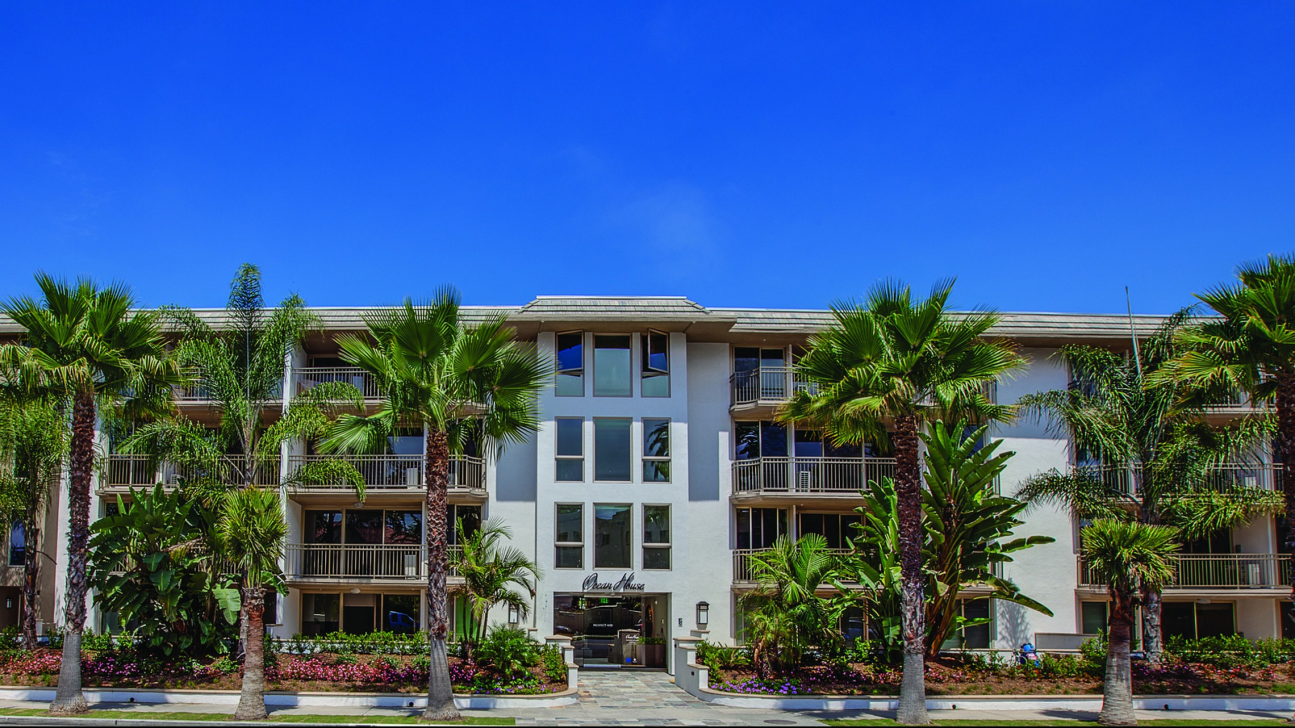 Elegant apartment building with lush palm trees, stunning architecture, and vibrant landscaping under a clear blue sky.