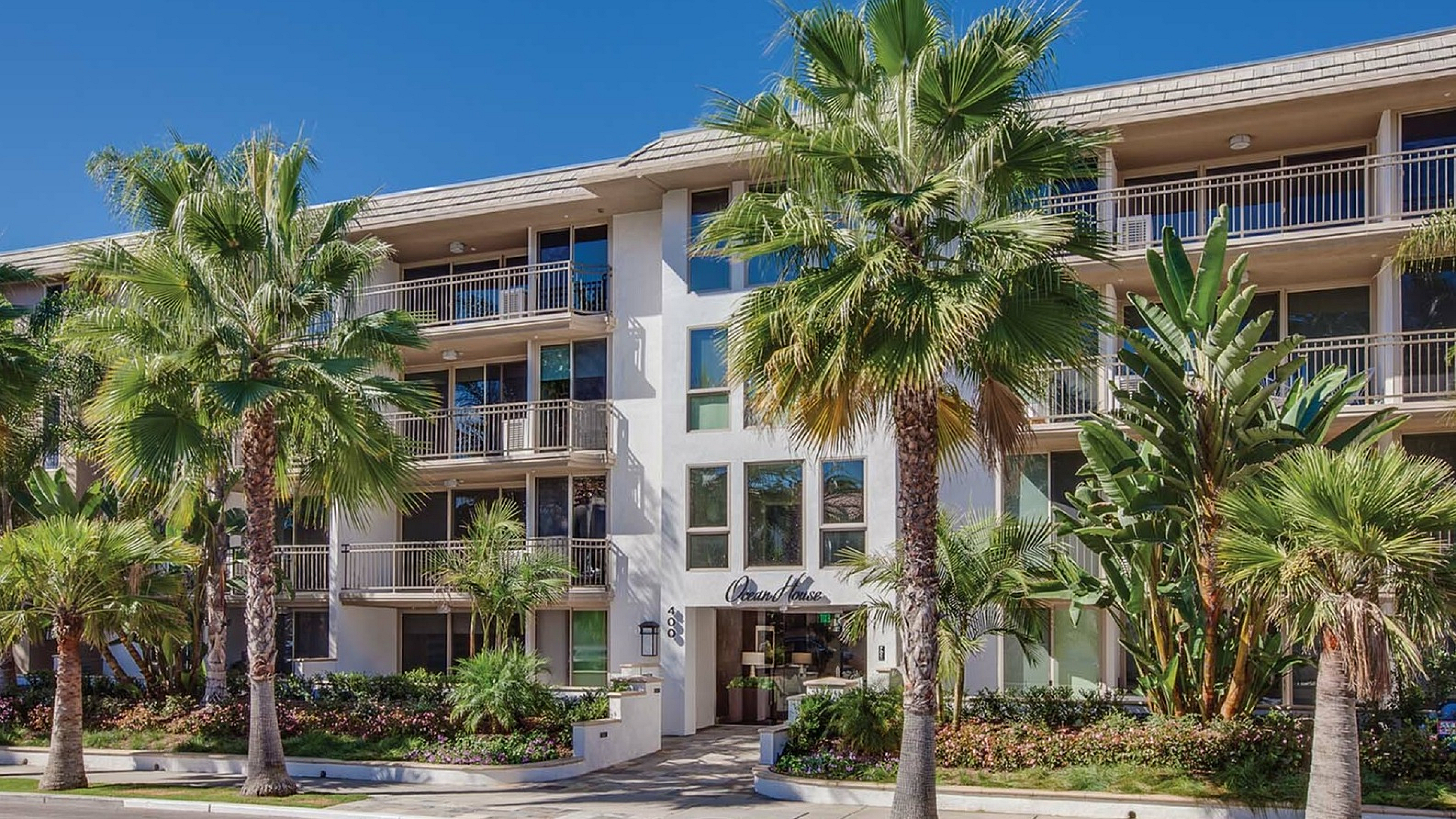 Beautiful apartment building with elegant balconies surrounded by lush palm trees and vibrant landscaping under a clear blue sky.