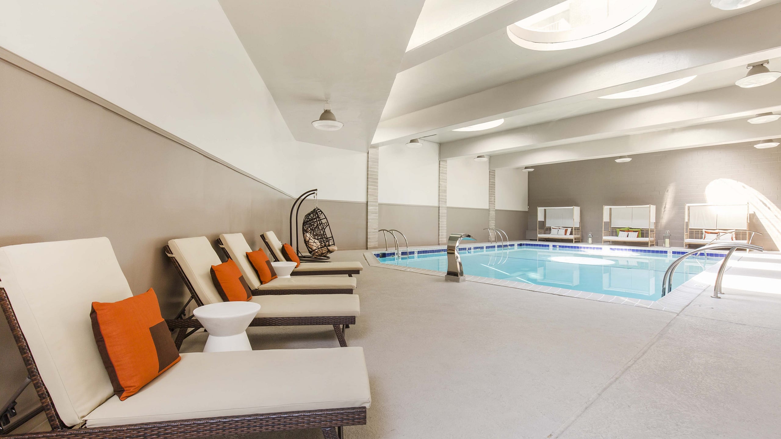 Luxurious indoor swimming pool area with elegant lounge chairs, vibrant orange accents, and skylight design for natural illumination.