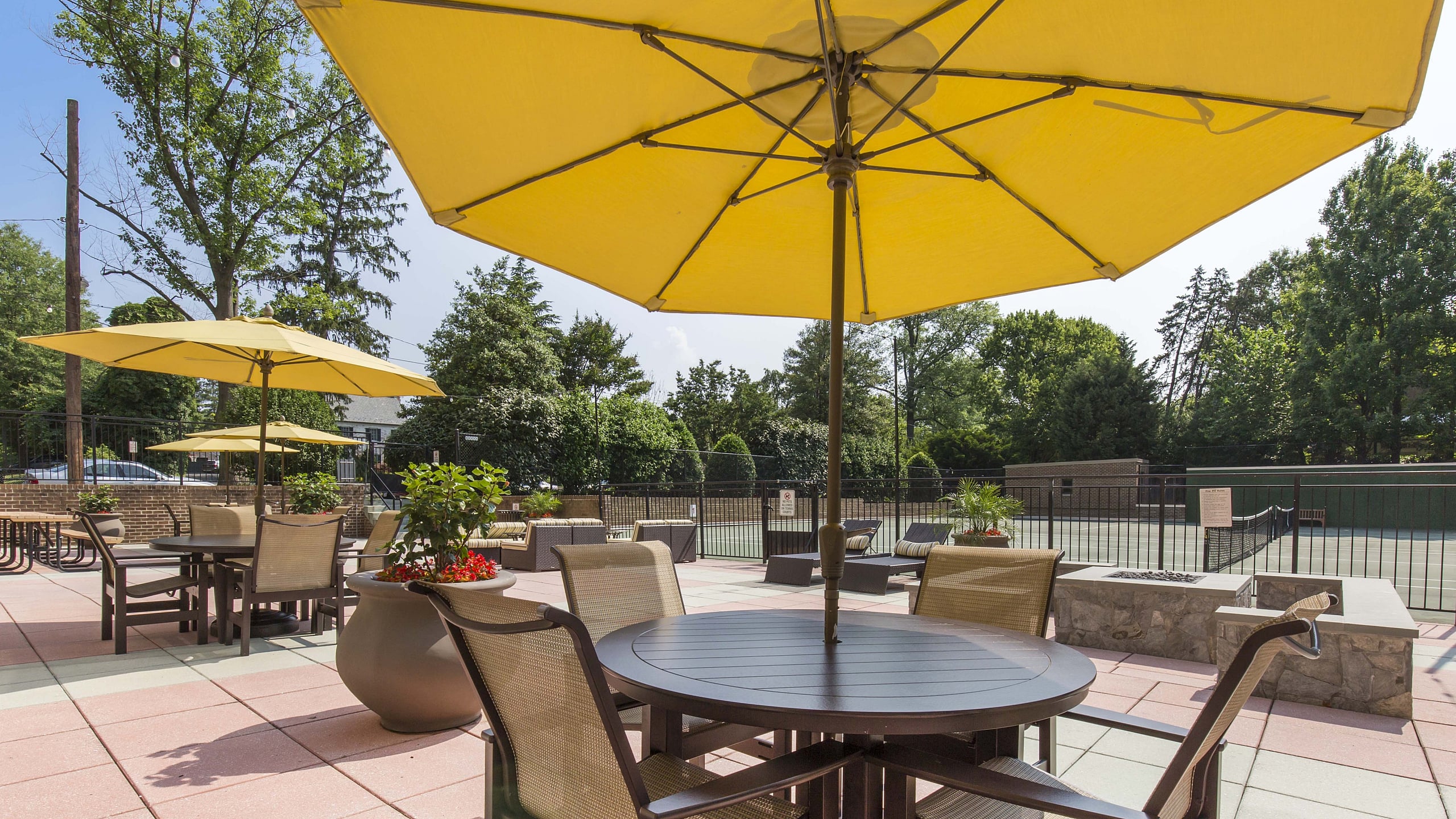 Vibrant outdoor patio with stylish seating under yellow umbrellas, lush greenery, and a tennis court backdrop.