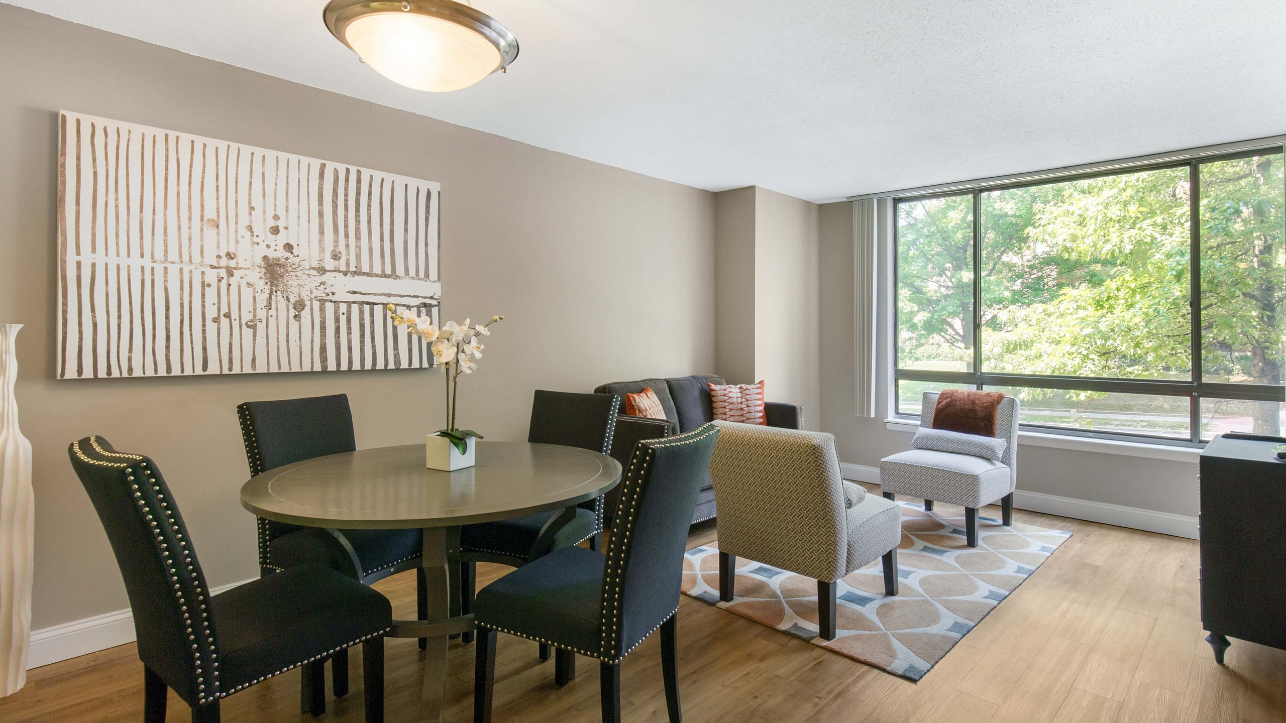 Bright and inviting apartment living area with stylish decor, featuring a cozy seating arrangement and a dining area under elegant lighting, set against large sunlit windows and warm wooden flooring.
