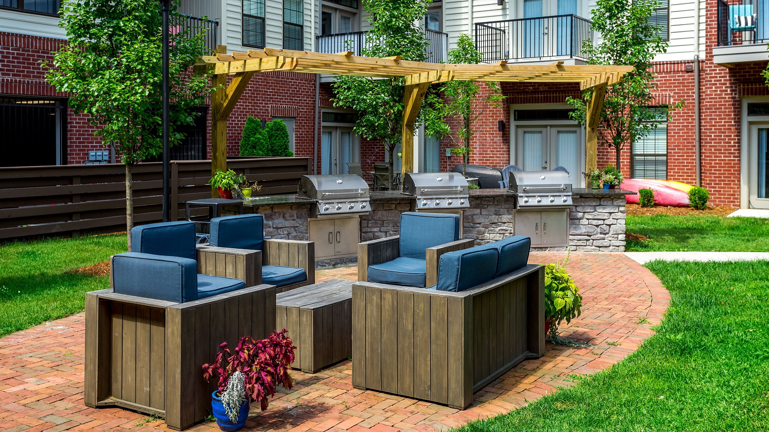 Inviting outdoor patio in modern apartment complex with stylish seating, lush greenery, and stainless steel grills under a charming pergola.