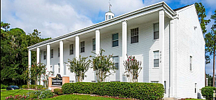 Elegant white colonial-style apartment building with grand columns and lush green landscaping under a bright sky.