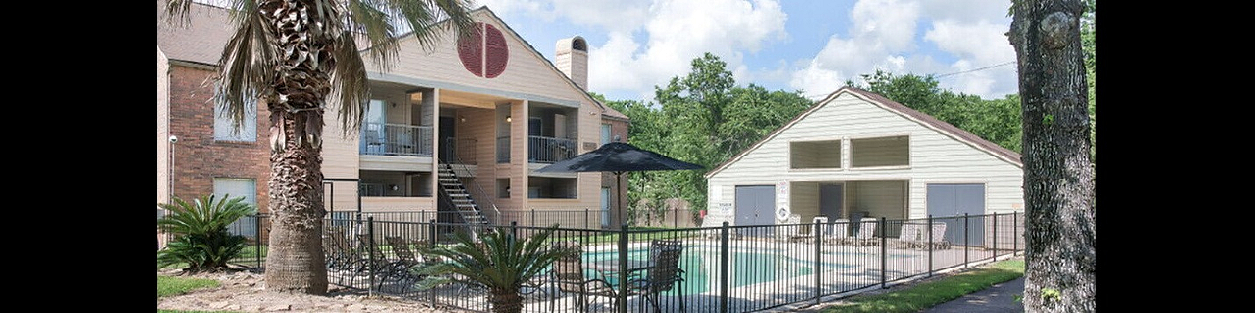 Charming apartment exterior with lush landscaping, palm trees, and a sparkling pool surrounded by a stylish iron fence.