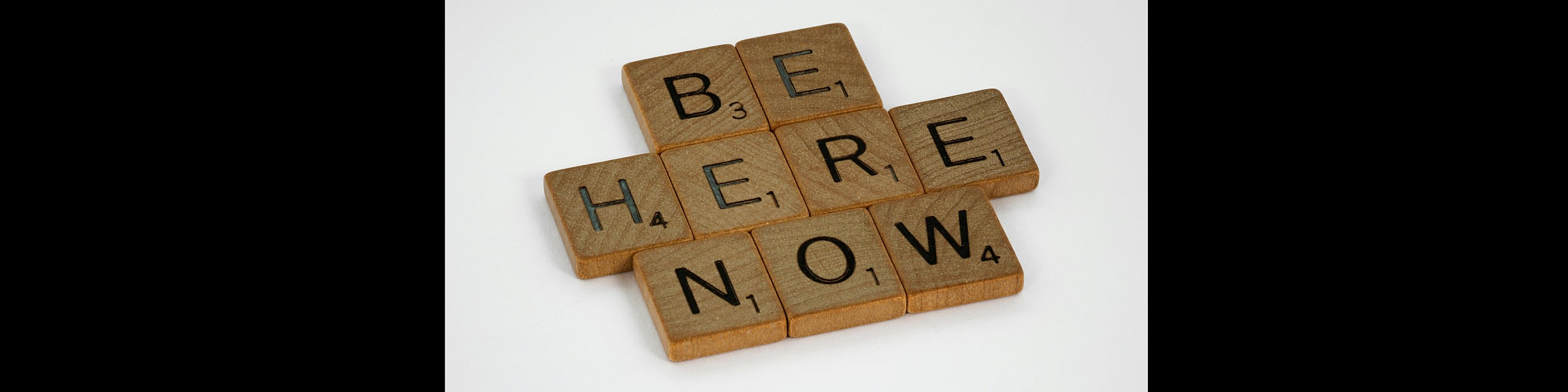 Inspiring word art formed by scrabble tiles spelling 'BE HERE NOW', emphasizing mindfulness and presence.