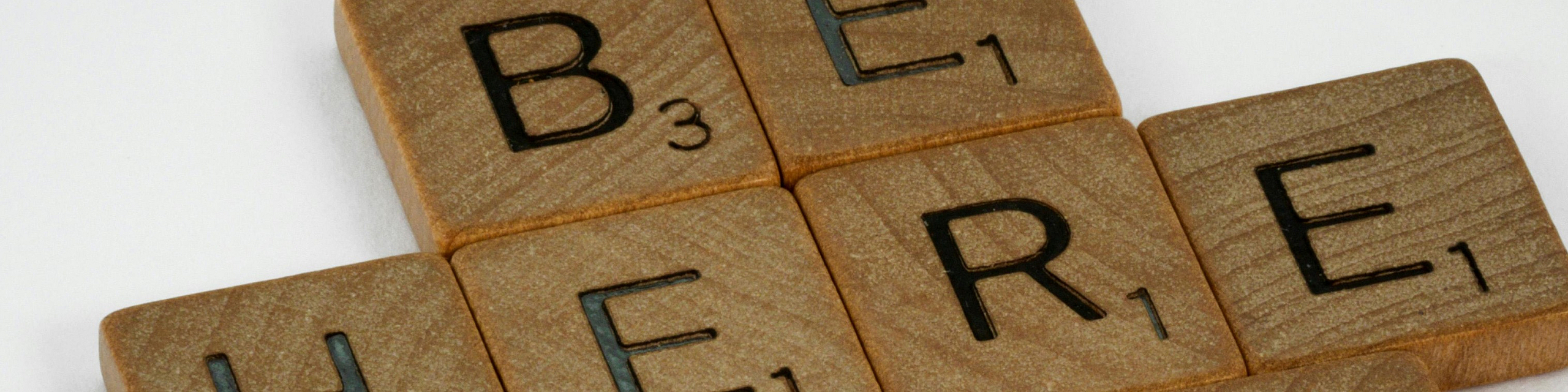 Inspirational message displayed using wooden Scrabble tiles forming the phrase 'Be Here Now' on a clean white background.