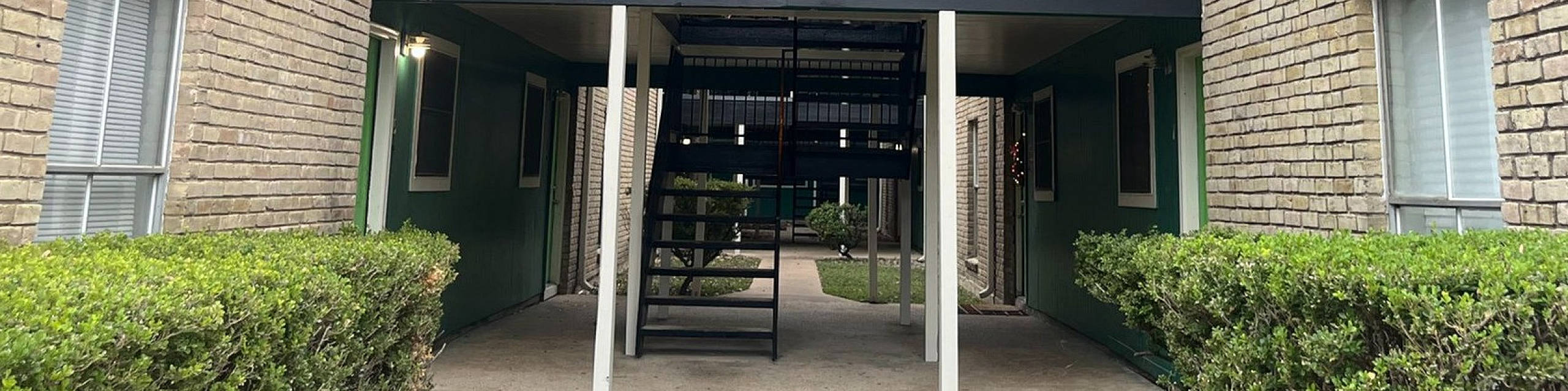 Charming apartment building entrance with classic brick exterior, cozy patio, and lush greenery, offering a welcoming vibe.